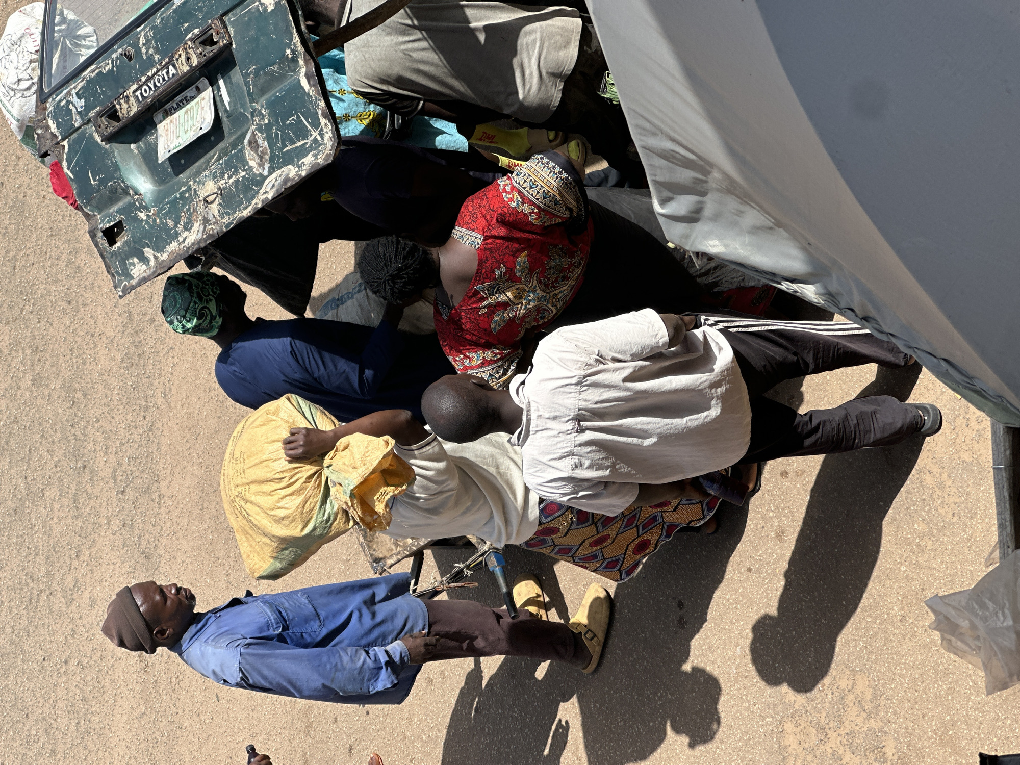 In Bukuru, Nigeria, a group of individuals are engaged in loading a large, dented green car door onto a truck. A man wearing a blue jumpsuit and a beanie stands on the left, looking towards the activity. Next to him, a person in a white shirt and patterned skirt carries a large yellow sack on their head. To the right, a person in a blue shirt and a green patterned head covering stands near the car door. Another person, seen from behind, wears a red and black patterned top and black pants. A young person in a light-colored shirt and black pants is also present. The environment appears to be a dusty, outdoor area, possibly a market or roadside, with a paved ground surface. The car door is prominently displayed, showing the "TOYOTA" logo and a license plate. The overall scene suggests a communal effort in transporting goods or salvaged materials.