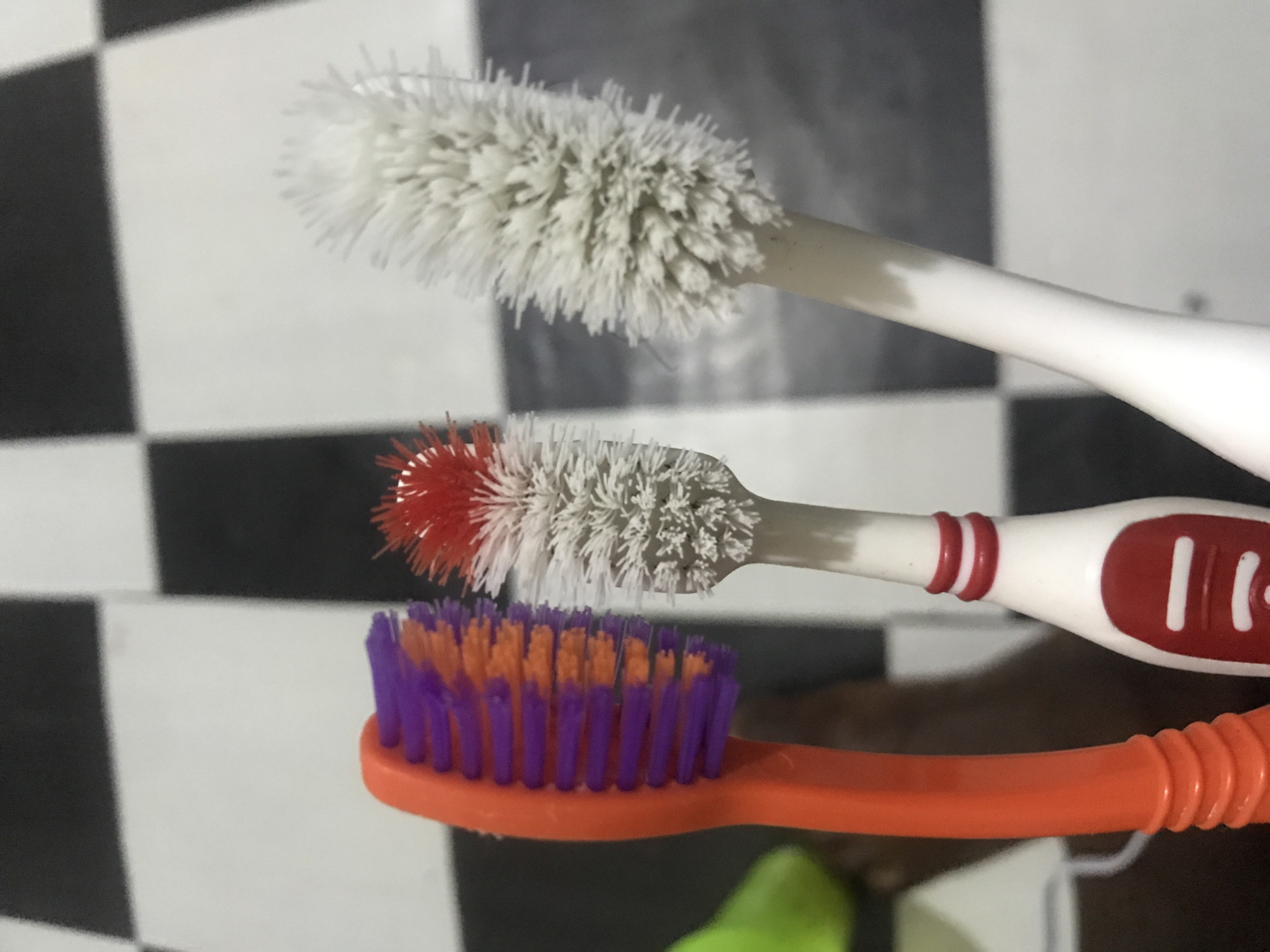 A close-up, eye-level, medium shot shows three toothbrushes with worn bristles. On the left, an orange-handled toothbrush with purple and orange bristles is angled towards the right. To its right, a white toothbrush with a red band and a red oval inset has red and white bristles that are splayed and worn. To the right of that, a white toothbrush with white bristles that are extremely worn and frayed is angled to the left. The background consists of a black and white checkered floor. The scene is indoors, with no discernable location within a city.