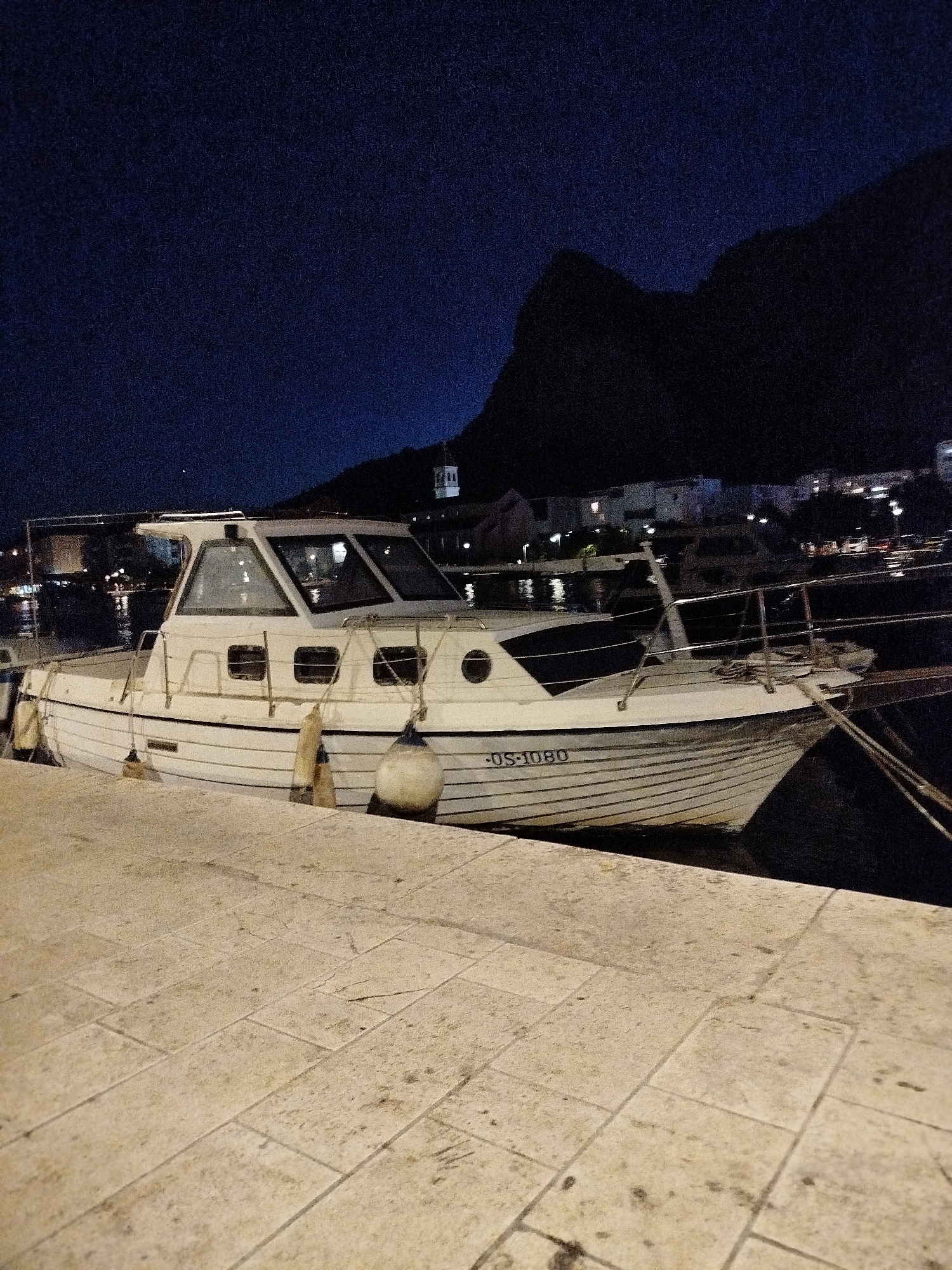 A white motorboat is docked at night along a paved waterfront in Omiš, Croatia. The boat, which has the registration DS-1080 visible on its hull, is white with black trim and windows. It is secured by ropes to the dock and has several buoys attached. In the background, lights from buildings and the dark silhouette of a mountain range are visible. The sky is dark blue.