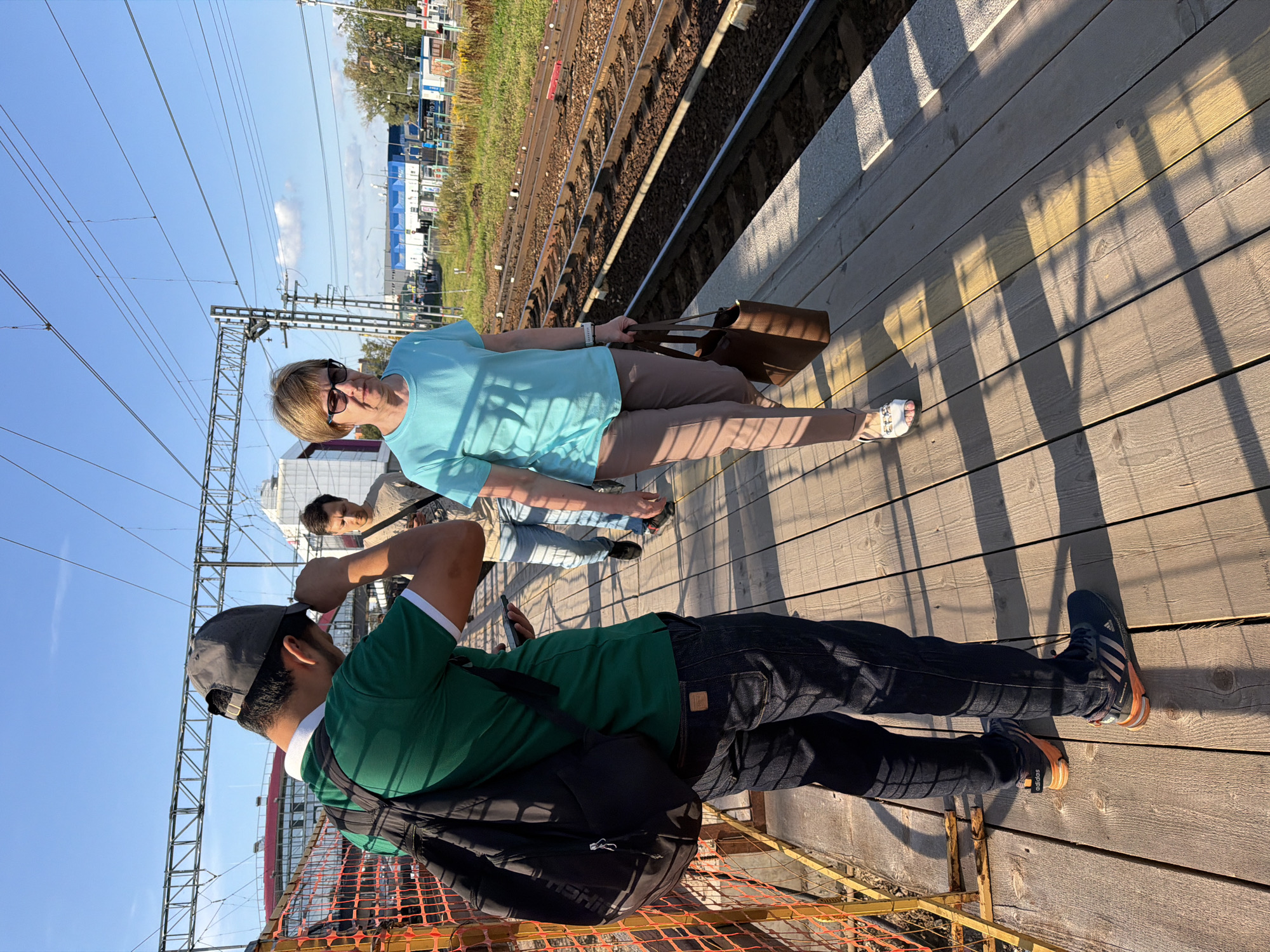 The image captures a daytime scene at a train station. In the foreground, a person wearing a green t-shirt, dark jeans, and a black backpack is standing on a wooden platform, adjusting their baseball cap and holding a smartphone. To their right, a woman with short blonde hair, wearing sunglasses, a light blue t-shirt, and taupe pants, walks away from the camera, carrying a brown tote bag. Behind them, another person in a beige t-shirt and jeans is walking further down the platform. The environment includes train tracks running alongside the platform, overhead electrical wires, and various industrial or commercial buildings in the background under a clear blue sky with some scattered clouds. The sun casts long shadows on the wooden platform.