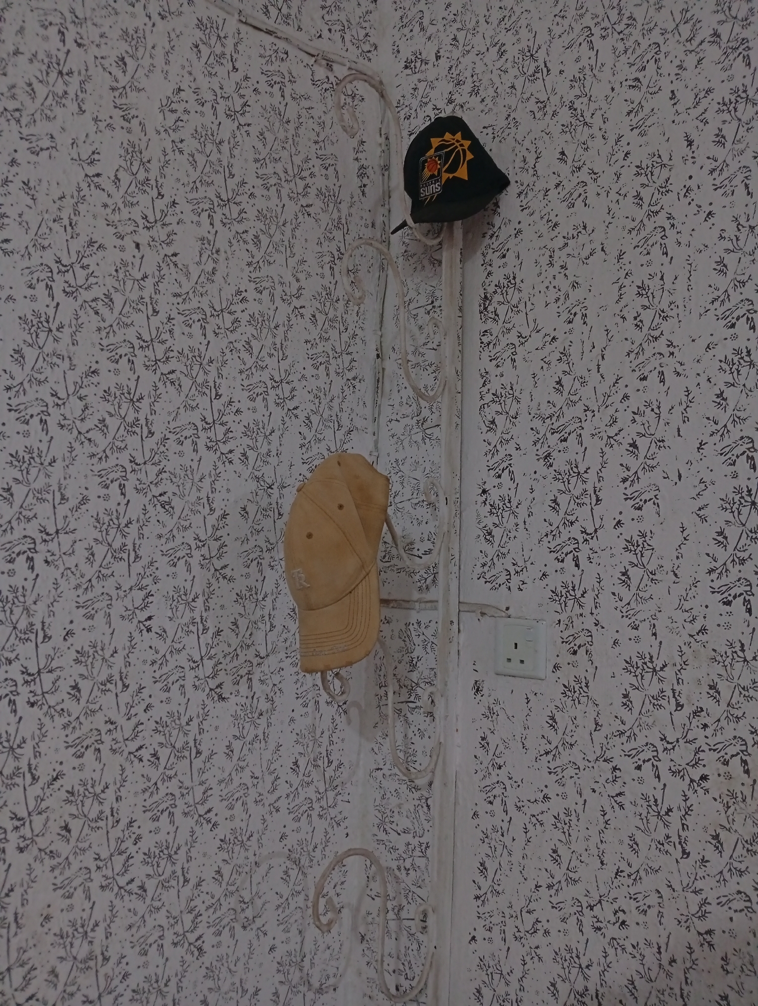 A photograph shows two baseball caps on a white metal coat rack in a room in Lokoja, Nigeria. The room has patterned white wallpaper. The coat rack is mounted on the wall, next to a white electrical outlet. One cap is black with a yellow and orange logo, and the other is beige.