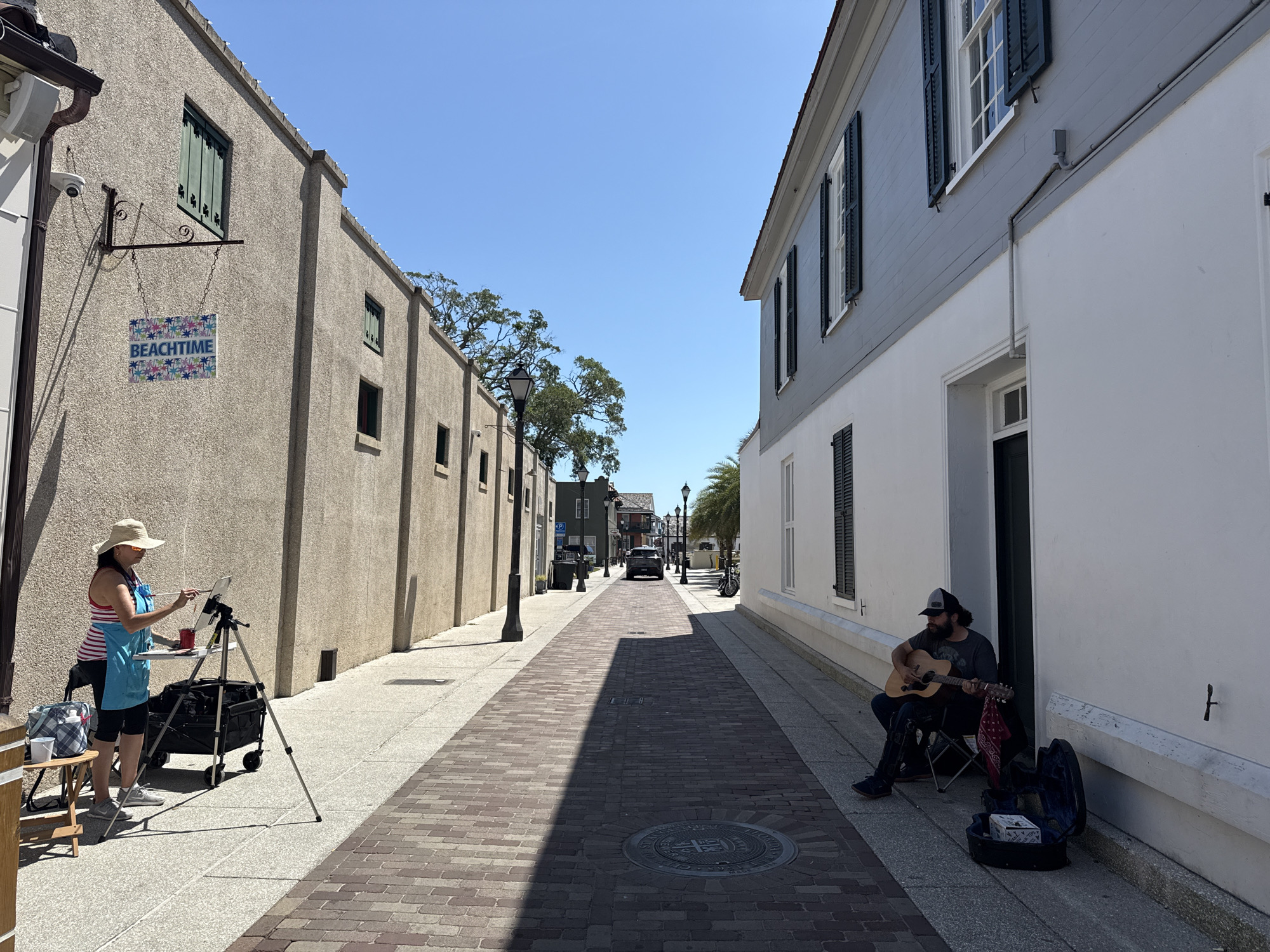 An outdoor scene on a brick-paved street in Saint Augustine, Florida. An artist is painting on a tripod on the left. The building has a "Beach Time" sign on its wall. A musician is playing guitar, seated on the right. Buildings with windows and shutters line both sides of the street. Street lamps are visible. A vehicle is down the road. The sky is clear and blue.