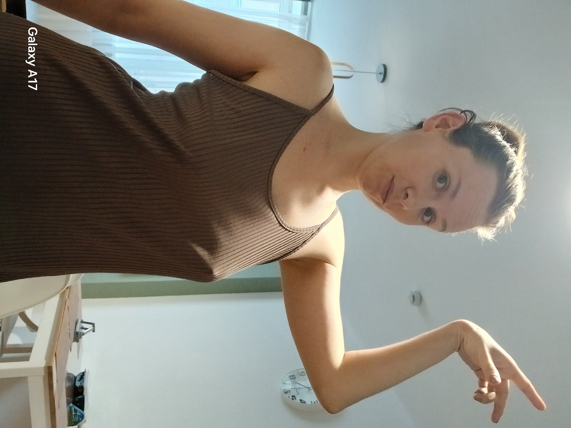 A woman in a brown tank dress is posing for a picture. She is pointing downwards. A white round clock is seen on the wall. A white table and chair can be seen. The location is not mentioned in the image, therefore, it cannot be determined.