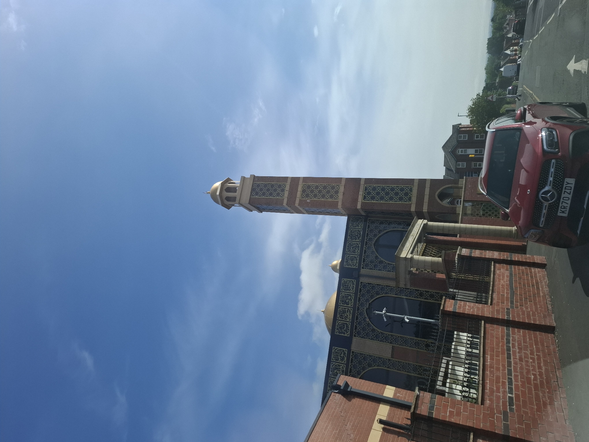 A wide shot captures the exterior of a mosque in Manchester, United Kingdom, on a bright, sunny day with a blue sky dotted with sparse white clouds. The mosque features a tall, brick minaret topped with a golden dome, and a main building with a large golden dome. The architecture incorporates intricate golden Islamic calligraphy and geometric patterns against a dark facade, with large arched windows. In the foreground, a red Mercedes-Benz SUV is parked on the right side of the road. The vehicle has the license plate KR70 ZDY. A red brick wall with a black metal fence lines the front of the mosque. To the right of the mosque, a residential street with houses and trees extends into the distance. The road has white lane markings and a pedestrian crossing.