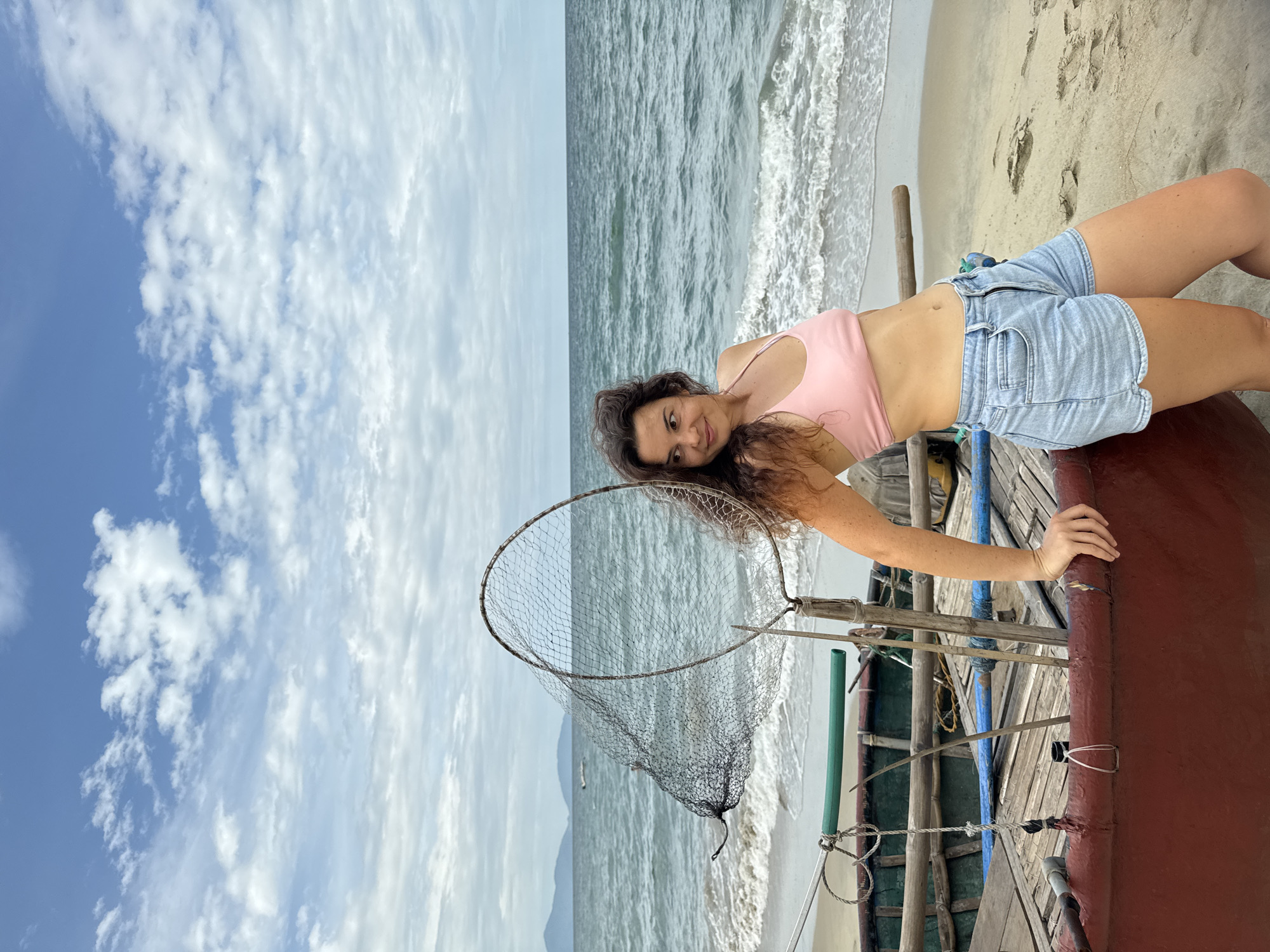 A young woman stands leaning on a traditional Vietnamese fishing boat on a sandy beach. She is wearing a pink bikini top and denim shorts. The sea is calm with gentle waves, and a net is propped up on the boat. In the background, a hazy coastline with mountains is visible under a partly cloudy sky. The scene appears to be An Bang Beach in Hoi An, Vietnam, known for its fishing boats and relaxed atmosphere.
