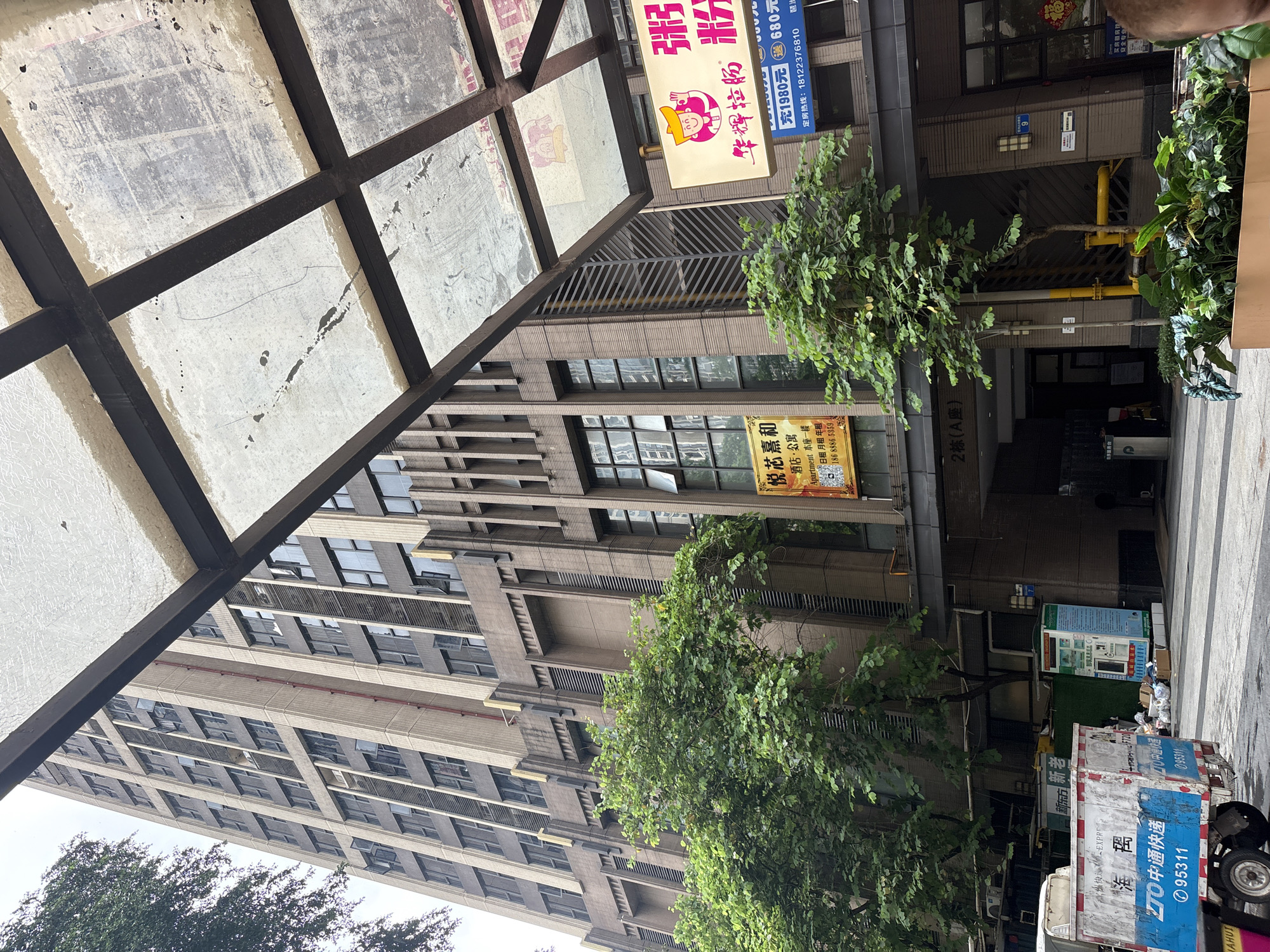 This is a medium shot, eye-level outdoor photograph taken from a street in Haizhu District, China, looking up at a tall, modern apartment building. The building's facade is a mix of beige and dark gray materials, with numerous windows and vertical architectural elements. A covered walkway with a dirty, translucent roof and a dark metal frame is in the foreground, partially obscuring the view of the building.
There are several signs on the building. One is a yellow rectangular sign with Chinese characters and an image of a building. Another, to its right, is a lit, rectangular sign with a cartoon character and Chinese text. Below this, a smaller blue sign has Chinese text and numbers. A tree with green foliage is growing in front of the building, partially obscuring some of the lower levels.
To the left of the building entrance, there is a white and blue utility truck or garbage bin with Chinese text and numbers. A blue and white vending machine is visible further to the left. In the lower right corner, a person's shoulder and the back of their head are visible, suggesting they are either the photographer or someone standing nearby. The ground appears to be paved. The sky is overcast.