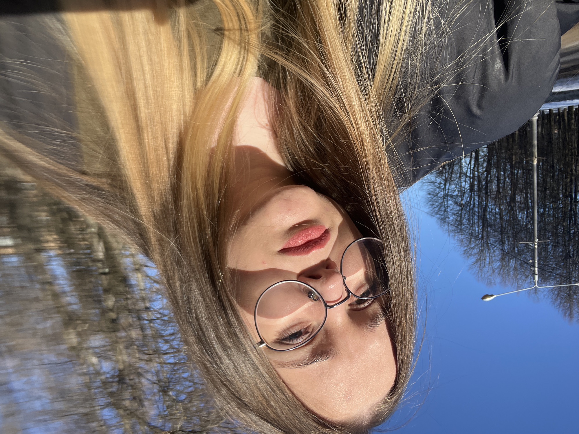 A young adult female is depicted in a close-up, selfie-style portrait taken outdoors in Pushchino, Russia. She wears round, metallic-framed glasses and reddish-pink lipstick, gazing directly into the camera. Her long, straight, light-colored hair, transitioning from brown at the roots to blonde ends, frames her face and extends over her dark-colored jacket and visible olive-green undershirt. Bright sunlight illuminates her from the left, creating strong shadows from her glasses on her face. The background, largely blurred, shows a clear blue sky above a line of leafless trees, consistent with late autumn or winter conditions. A street lamp on a tall pole is visible to the left, adjacent to a paved area with remnants of snow on the ground.