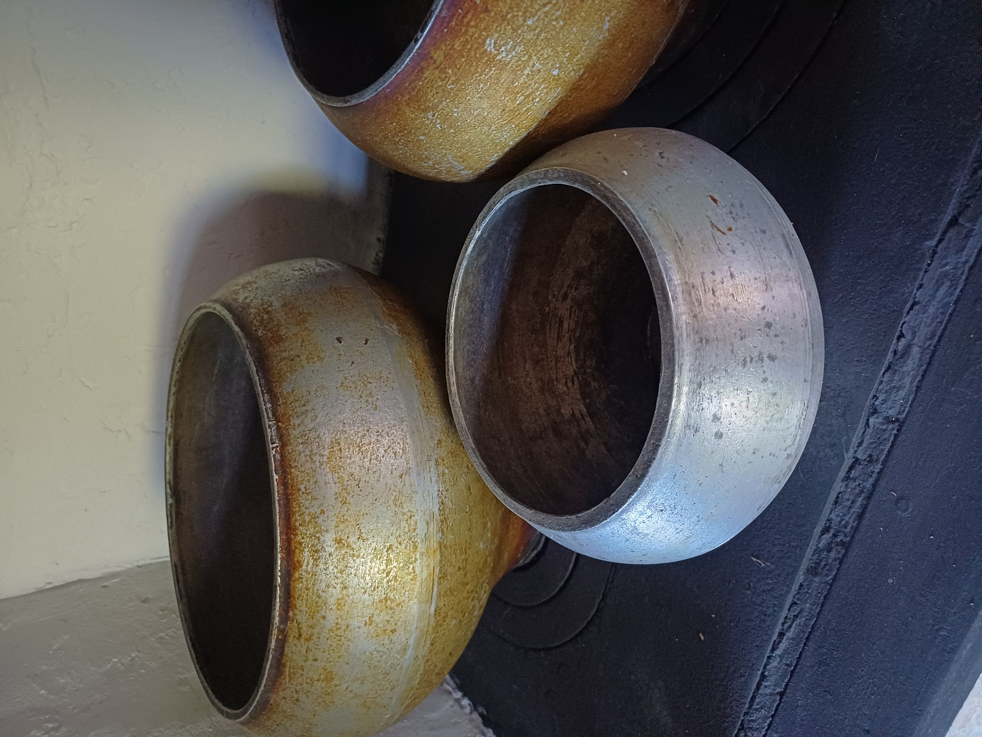 The image shows three metal pots with weathered, burnished surfaces, placed on a dark, rough surface, likely a stove top. The pots vary in size and color, with one in the foreground appearing silver, while the other two are a golden-brown hue with darker patches from heat exposure. The background is a textured, off-white wall. The scene depicts a rustic, domestic setting. The specific location of Sulymivka, Ukraine, is a detail that cannot be visually determined from the provided image alone.