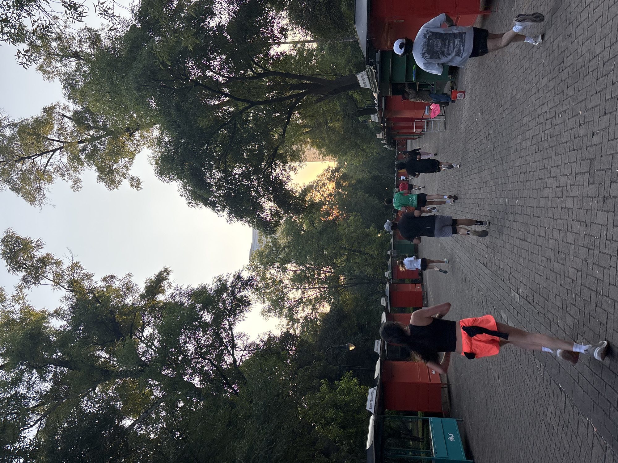 The image depicts a group of people running along a brick-paved pathway in Mexico City, Mexico. Trees line both sides of the pathway, creating a canopy overhead. The runners are wearing various athletic outfits, including shorts, t-shirts, and sneakers. The scene is bathed in the soft light of the setting sun, with glimpses of sunlight filtering through the trees. Red and teal structures are present on the side of the pathway.