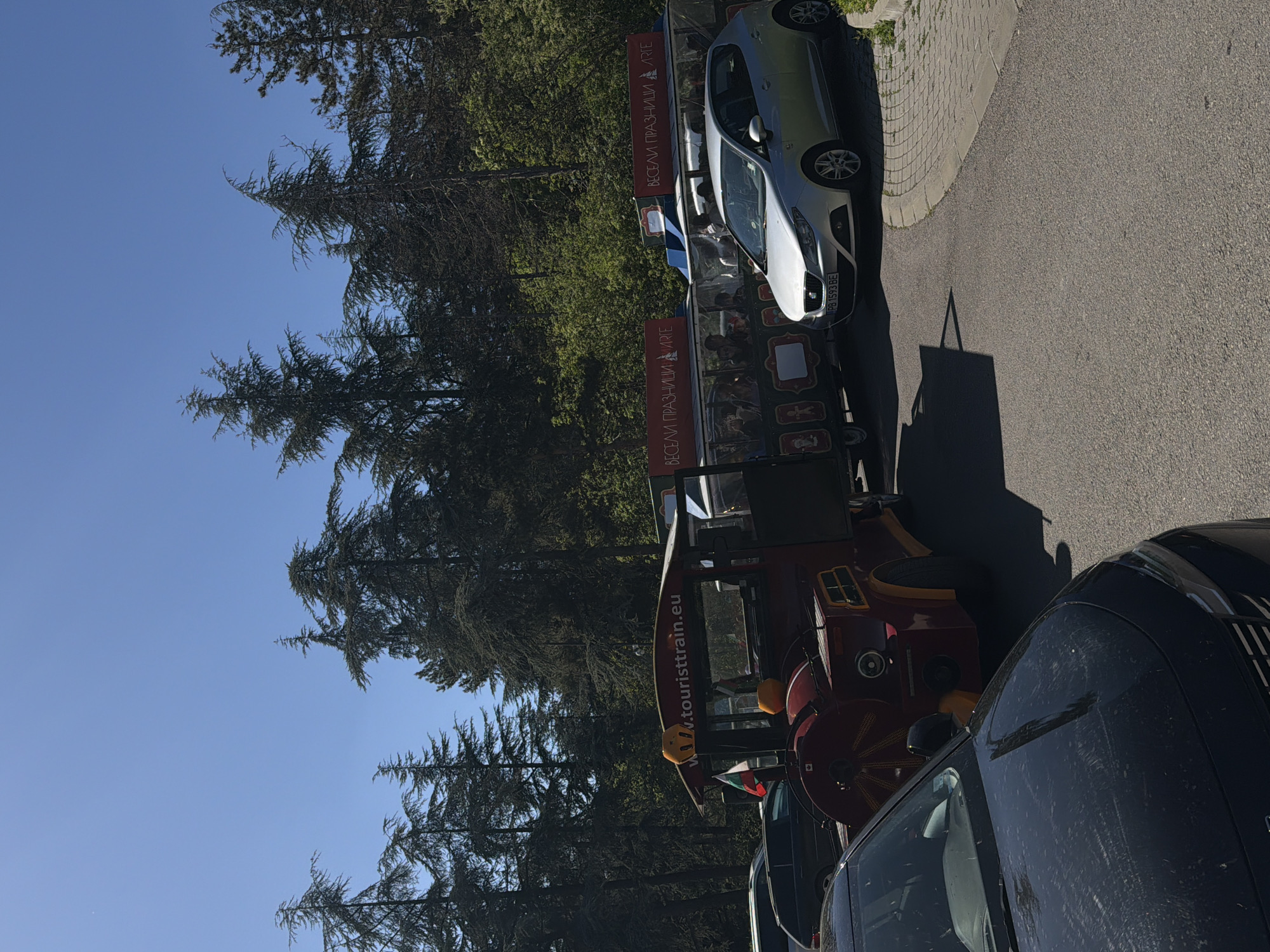 The image captures a street scene in Stara Zagora, Bulgaria. A red tourist train, labeled "touristtrain.eu" and adorned with the text "ВЕСЕЛИ ПРАЗНИЦИ ЛЯТО" (Happy Holidays Summer), stands parked near several cars. A silver car is positioned partially on the sidewalk. A black car is parked at the front of the tourist train. The trees on the left side of the image suggest the location is near a park or green area. The clear blue sky indicates it is a sunny day.