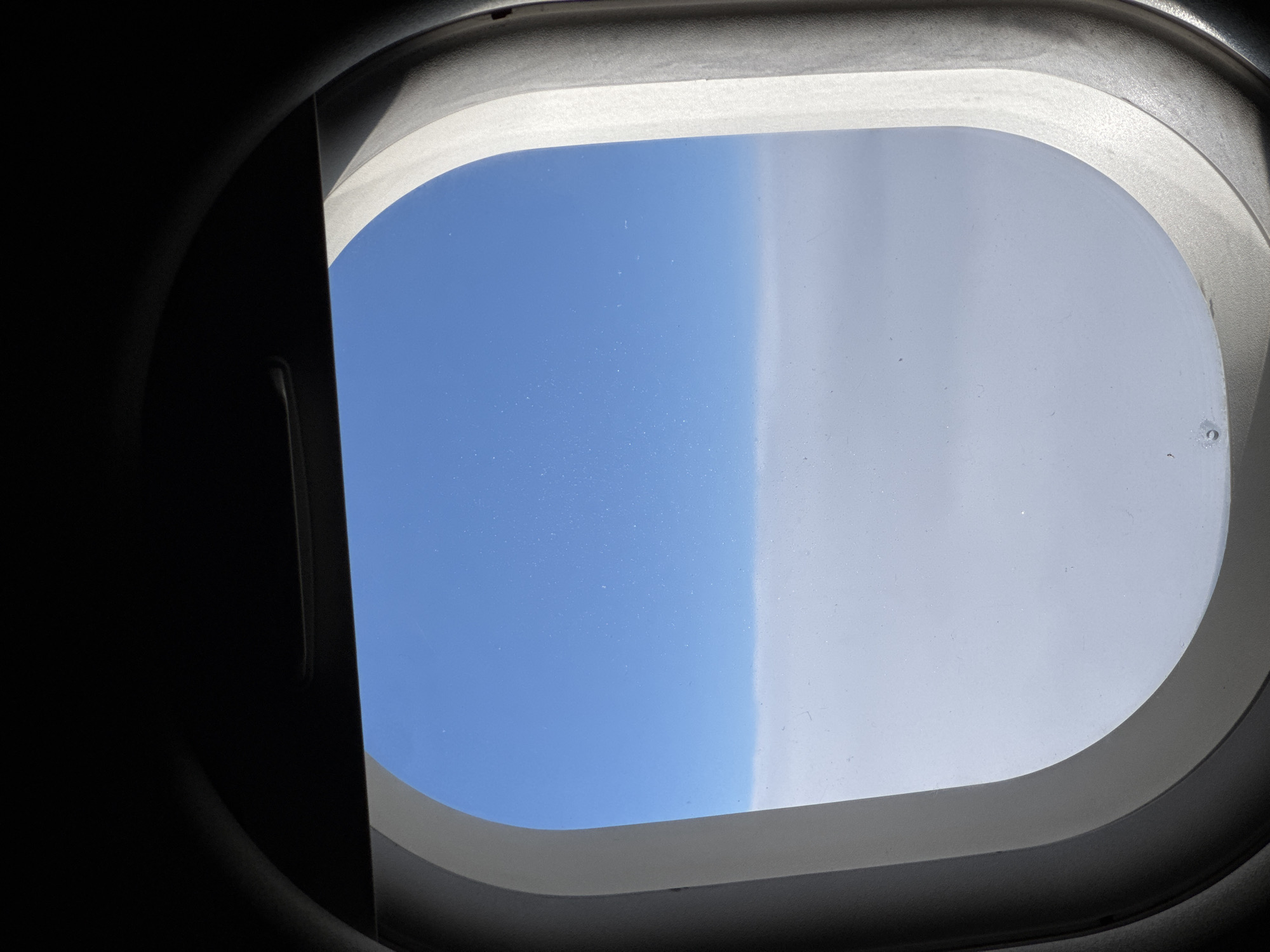 The image is a view from an airplane window. The window frame, which is dark on the top and a light, textured gray on the sides, encloses a view of the sky and clouds. The sky is a clear, bright blue, with a few faint white speckles, possibly distant contrails or atmospheric particles. Below the blue sky is a thick layer of fluffy white clouds, creating a distinct horizontal line across the lower half of the frame. The perspective suggests the airplane is flying above the cloud layer, at a high altitude. The city of Ob, Russia, is not depicted in this image, as the scene is entirely comprised of sky and clouds viewed from an aircraft.