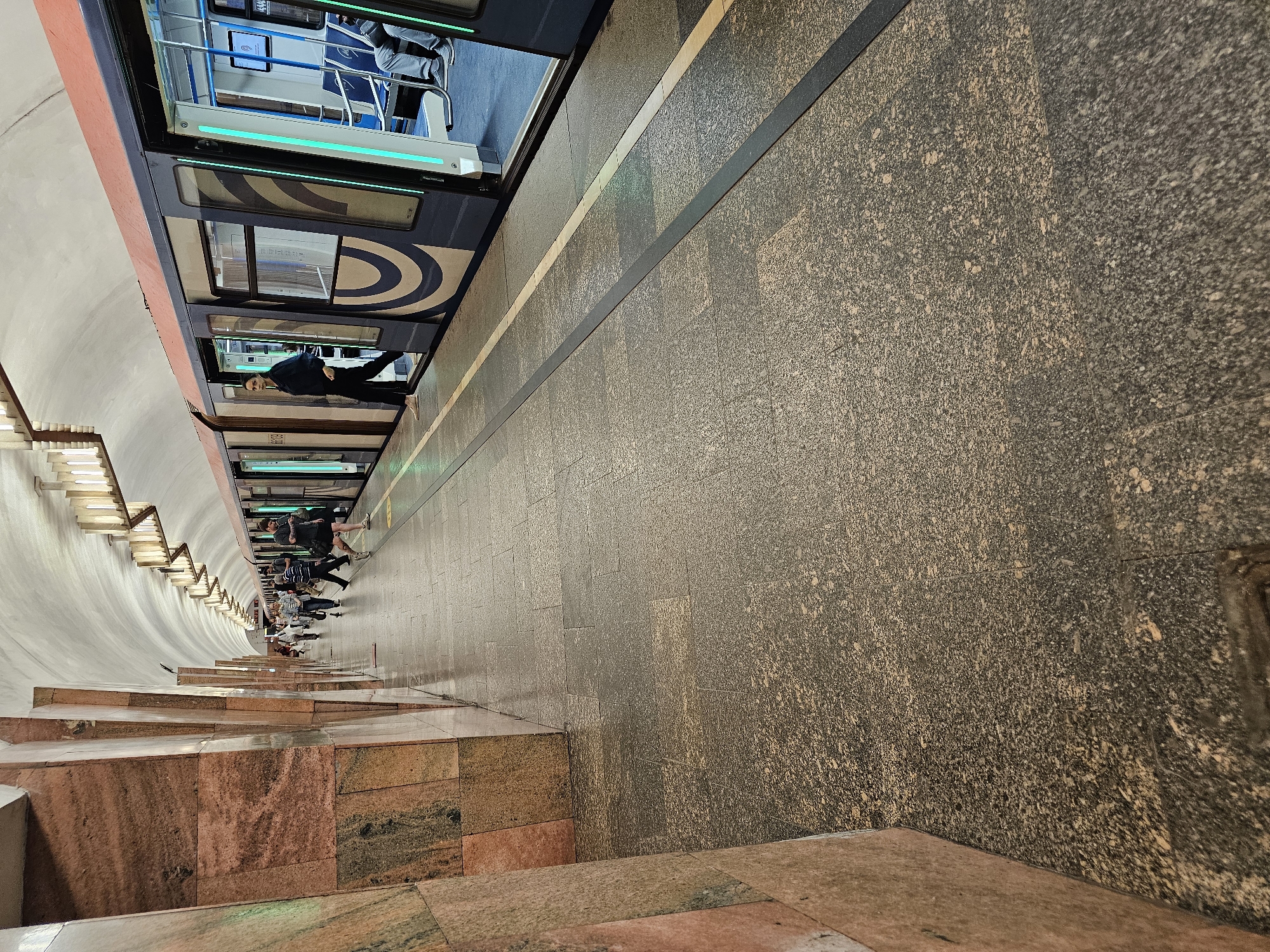 The image depicts a subway platform in Moscow, Russia. A modern blue and white train is stopped at the platform, doors open, allowing passengers to enter and exit. Several people are visible on the platform near the train, waiting to board or having just disembarked.
The platform itself is paved with patterned brown and grey stone tiles. A dark line runs parallel to the train, separating the platform edge from the walking area. The walls of the subway station are curved and covered with light-colored tiles. A decorative lighting structure lines the upper part of the wall.
In the foreground, stairs made of red and brown marble-like stone lead down to the platform level. The scene shows a typical moment of transit within the Moscow metro system.