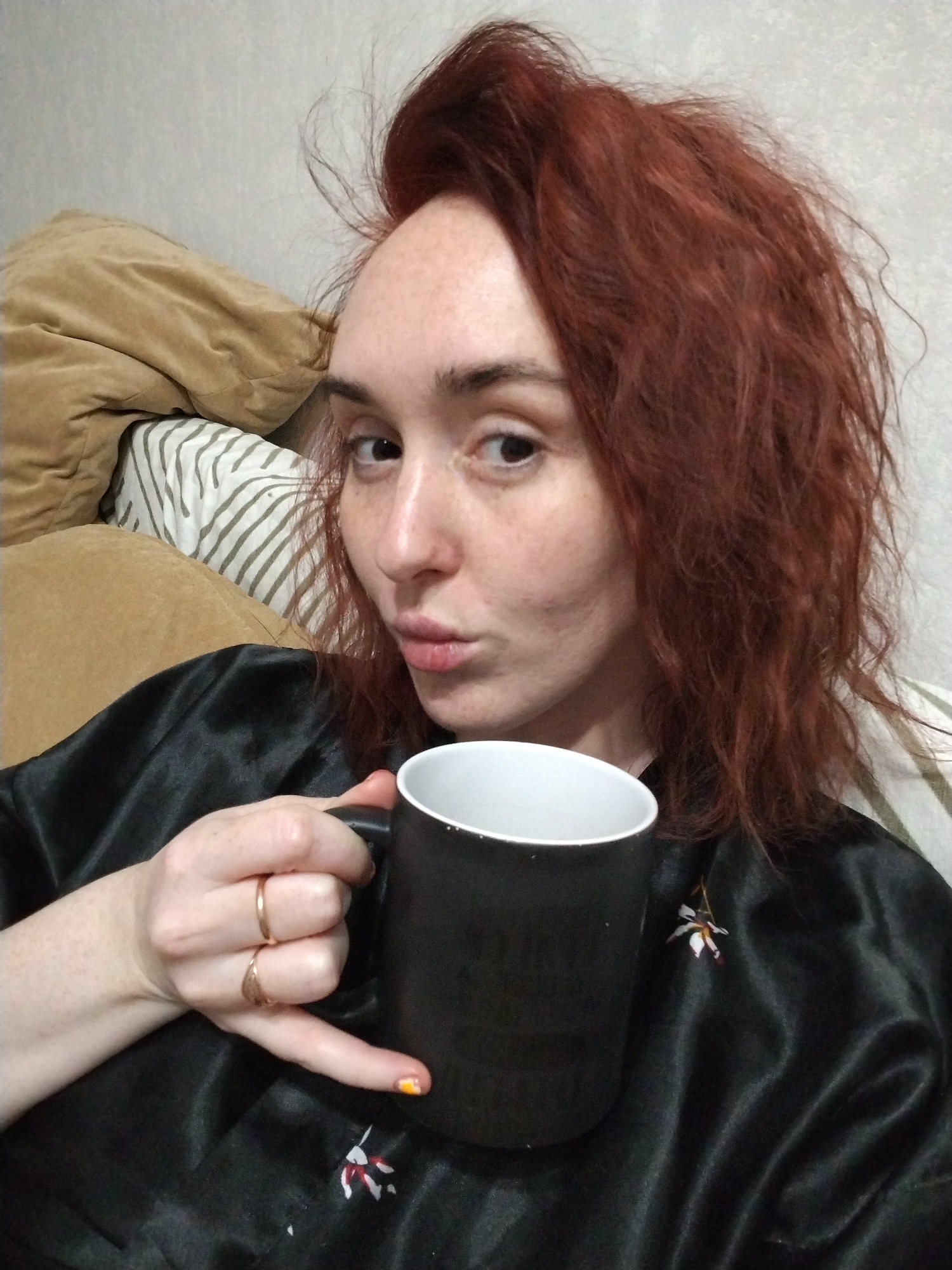 The image displays an adult female in an indoor setting, contextually placed in Izhevsk, Russia. She has reddish-brown, disheveled hair and a fair complexion with visible freckles across her face and on her left hand. Her lips are pursed, and she maintains direct eye contact with the camera. She is wearing a black, satin-like garment, which appears to be a robe or sleepwear, featuring a small, white and reddish-brown abstract or floral pattern on the right side of her chest. Her left hand, adorned with two gold-toned rings on the ring finger and an orange and yellow painted fingernail on the pinky, holds a black ceramic mug with a white interior. The background consists of a plain, light gray wall, a brown, plush textile object, and a white and dark-striped pillow or cushion, indicating a casual, domestic environment.