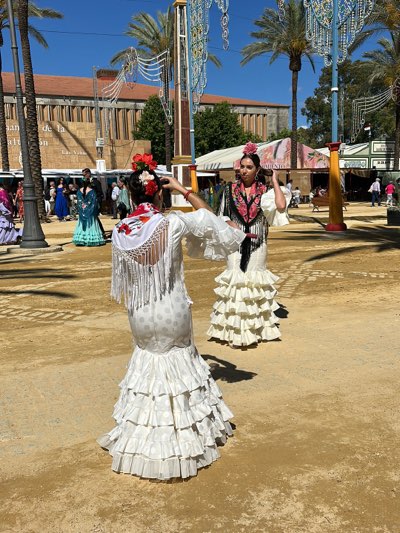 The image captures a scene at the Jerez de la Frontera fair in Spain. Two women are the primary subjects, both adorned in traditional flamenco dresses. One is turned away from the viewer, her white polka-dot dress detailed with ruffled layers at the skirt's base and a fringed shawl draping her back. Red flowers embellish her hair, and she is gesturing with her right arm. The other woman faces the viewer, wearing a similar white ruffled dress complemented by a black and red shawl and a flower in her hair. In the background, the fair setting is evident with light poles, palm trees, building facades, and other people. The ground is a light-colored dirt or sand, typical of fairgrounds.