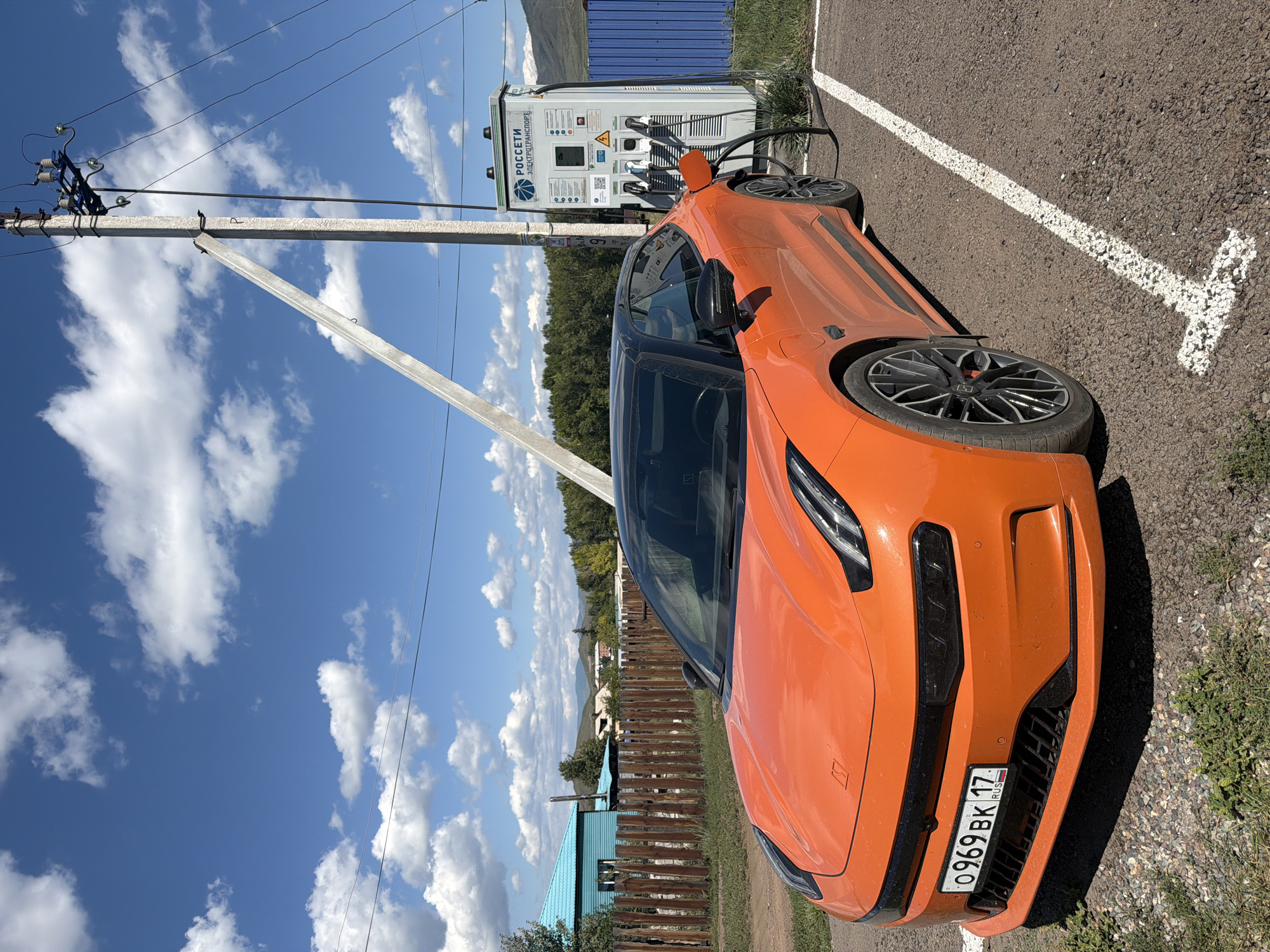 An orange electric car is parked next to a charging station in the city of Turan, Russia. The car is a coupe with a black roof and tinted windows. The charging station is white and has a blue logo with the words "РОССЕТИ ЭЛЕКТРОТРАНСПОРТ" written on it. The car's license plate reads "0969 BK 17". In the background, there are some houses, a wooden fence, and trees. The sky is blue with white clouds. The ground is covered with asphalt and gravel.