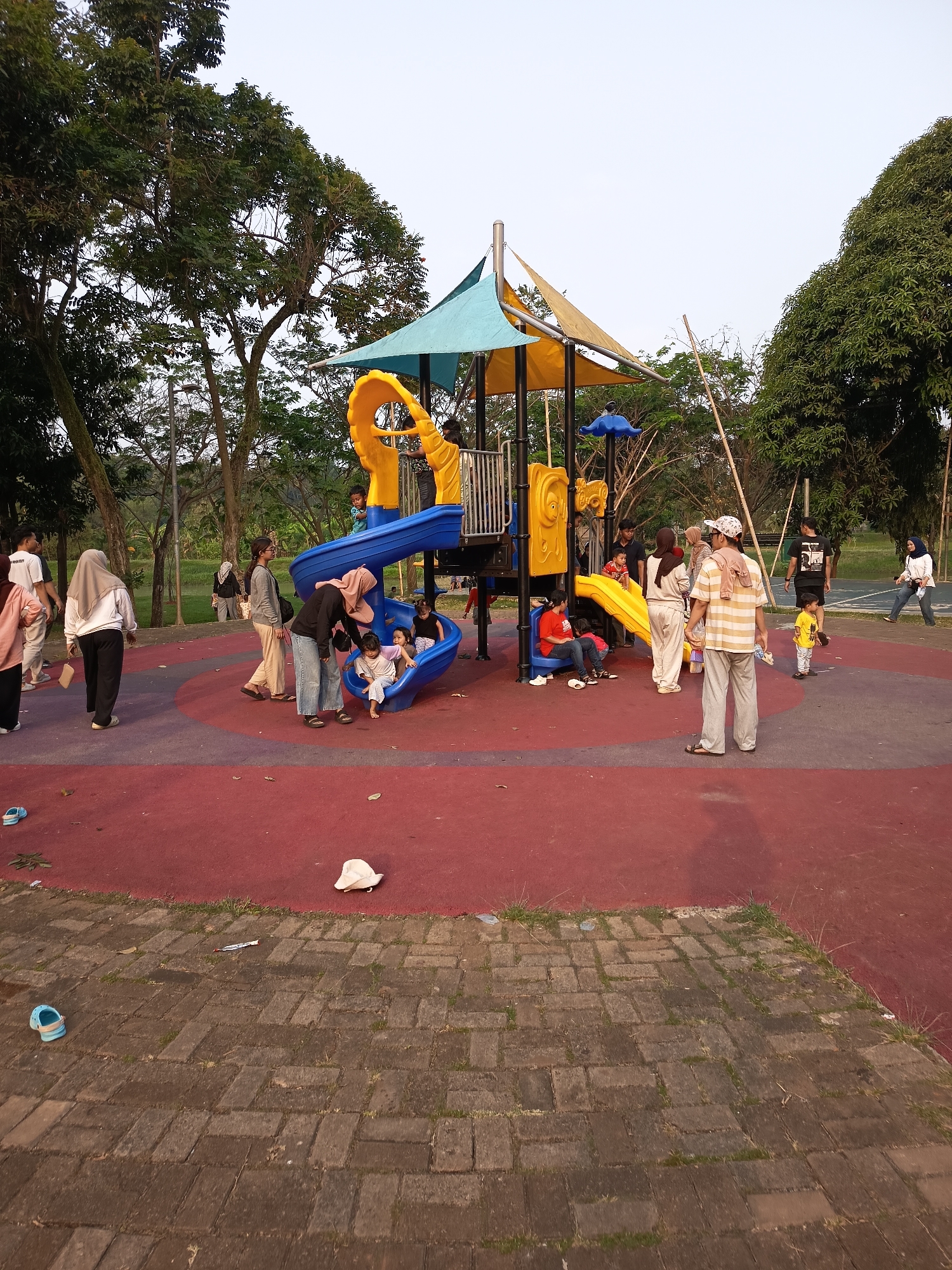 The image shows a playground in the city of East Jakarta, Indonesia. The playground features a multi-colored climbing structure with slides and a tunnel, a blue spiral slide, and a yellow slide. Several people, including adults and children, are present. Some are interacting with the playground equipment, while others are standing or walking around. The playground is situated on a red rubberized surface, surrounded by trees and a paved pathway. The sky is overcast.