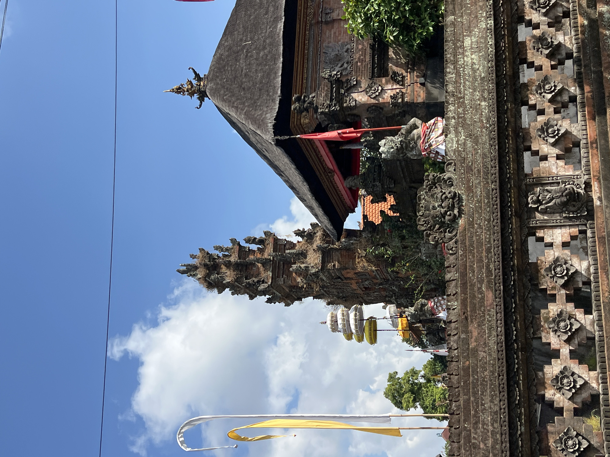 A temple complex is captured from a low-angle exterior view on a sunny day in Abiansemal, Indonesia. The foreground features a detailed stone wall with intricate carvings of floral patterns and mythological figures. Behind the wall, a multi-tiered pagoda-like structure is visible, adorned with yellow and white layered umbrellas. To the right, a larger temple building with a dark, thatched roof and brick walls stands prominently. Red and white flags are displayed on poles, and a single red flag is also attached to the main temple building. Two stone statues, one appearing to be a guardian figure with a patterned cloth around its waist, are positioned on the upper level of the temple wall. The sky above is bright blue with scattered white clouds. Tall trees and other temple structures are visible in the background.