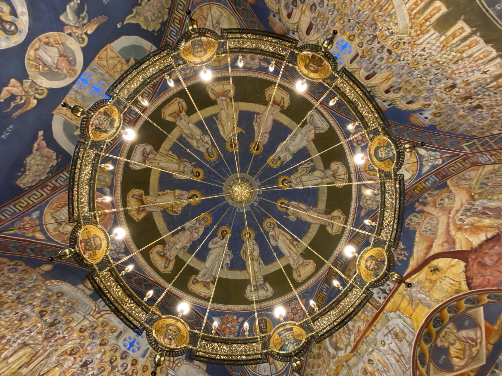 This image captures a low-angle, upward view of a magnificent, ornate chandelier suspended from the richly frescoed ceiling of a church or cathedral in Bar, Montenegro.
The central subject is a large, circular, multi-tiered chandelier, crafted from what appears to be gilded metal or brass, intricately decorated with scrolling patterns and small, illuminated light bulbs. Around the perimeter of the chandelier's main ring are several circular medallions, each containing a painted portrait of a bearded saint or religious figure, typically with a halo. Some visible text in Cyrillic or Greek script on these medallions identifies the depicted figures, for example, "СВ. ПЕТАР" (St. Peter) is discernible on one.
Above the chandelier, the domed ceiling is covered with vibrant frescoes in the Byzantine/Orthodox iconographic style. In the central circle of the dome, directly above the chandelier, a ring of robed, haloed figures, likely apostles or saints, stands on a dark blue background. Each figure is depicted standing on a raised platform, radiating outwards from the dome's apex. Further outwards, the ceiling features an expansive array of more haloed figures, often depicted in rows or clusters, representing a host of saints, martyrs, or angels. Geometric and floral patterns in red, gold, and blue adorn the structural elements of the ceiling.
The overall color palette is rich, dominated by deep blues, golds, earthy browns, and reds, creating a solemn and sacred atmosphere. The chandelier's bulbs are lit, casting a warm glow and highlighting the intricate details of both the metalwork and the ceiling art. No specific activity or event is taking place; the image is a static portrayal of the church's interior grandeur. The perspective is looking straight up, emphasizing the scale and detailed artistry of the ceiling and its central light fixture.
