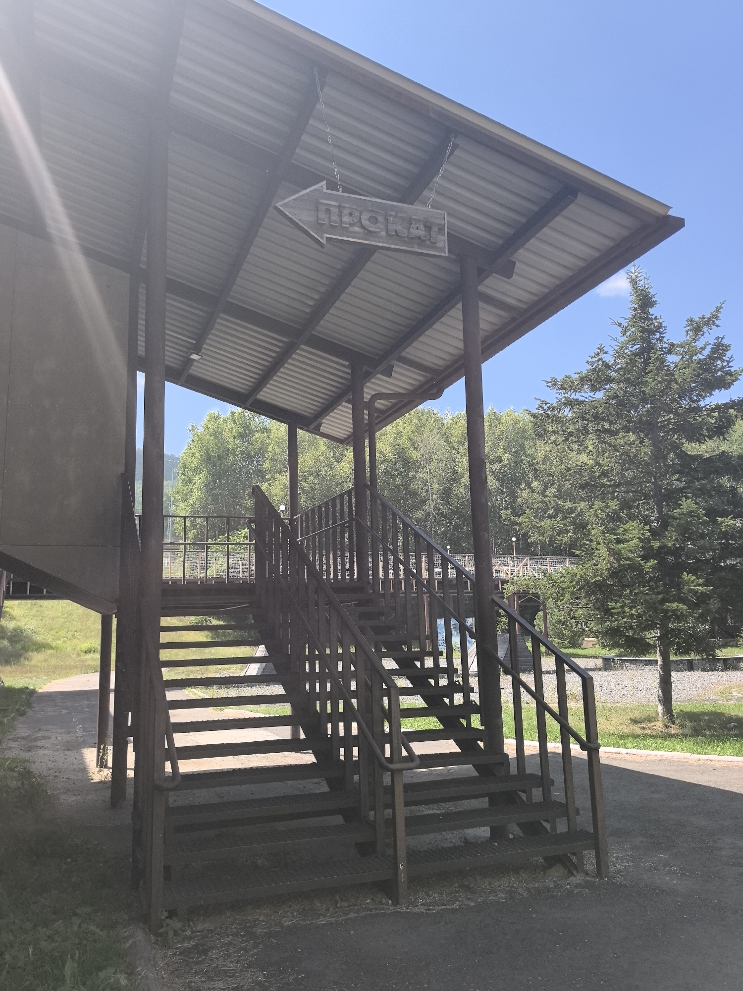 The image shows an outdoor stairway leading to a covered area with a sign that reads "ПРОКАТ" (PROKAT), which translates to "rental" in Russian. The sign is wooden and shaped like an arrow pointing to the left. The stairway is made of metal with a grated surface and is supported by dark metal posts. It is located in a park-like setting with trees and greenery visible in the background. The sky is clear and blue. The image appears to be taken in Solnechnyi, Russia.