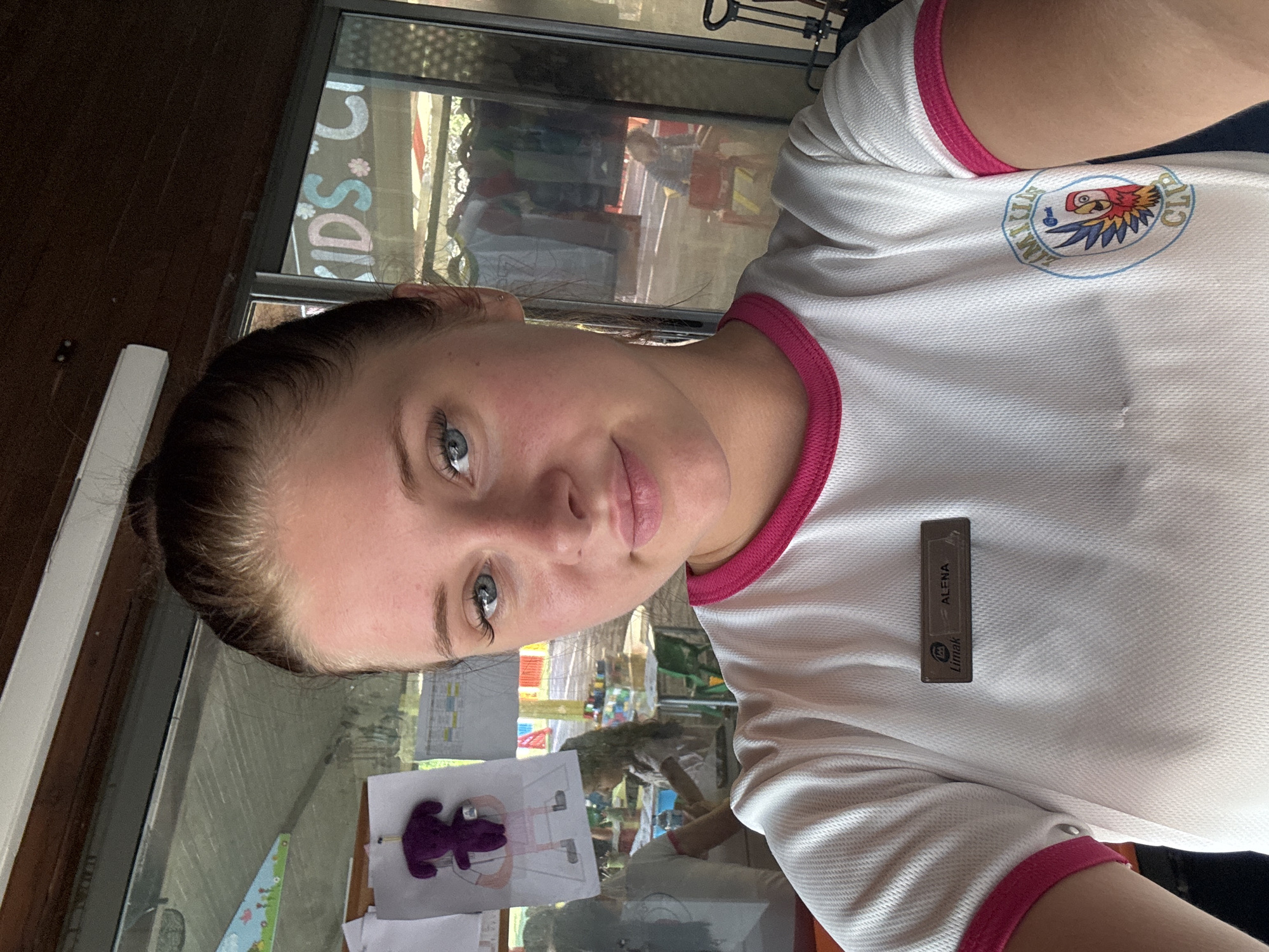 The image shows a young woman with her hair pulled back, taking a selfie indoors. She is wearing a white t-shirt with pink trim and a name tag that reads "Limak ALENA". A logo with a parrot and the words "Limak Club" is visible on the lower right side of her shirt. In the background, through a glass window, a room with tables, chairs, and children can be seen. A drawing of a purple stuffed animal is pinned to the wall. The setting appears to be a kids' club or daycare. The overall impression is a casual and bright environment, likely within the Limak Lara hotel in Aksu, Turkey, given the branding.