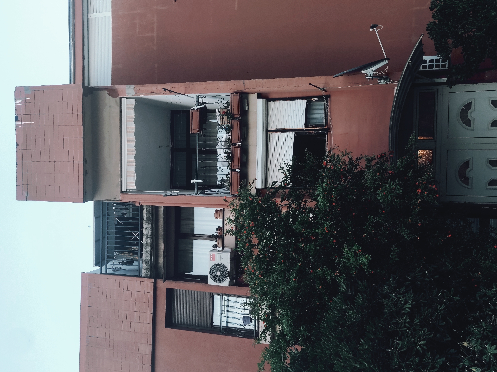 Here is a description of the image:
The image depicts the exterior of a multi-story building with a red-brown facade. Various windows are visible, some with open shutters and others with metal bars. An air conditioning unit is mounted on the building's exterior. A satellite dish is mounted on the wall near the top. A decorative tree or bush partially obscures the view, particularly near the bottom of the frame. A doorway with a curved overhanging structure is present. The location appears to be in an urban area, though the specific city and location are unknown. The sky is overcast.