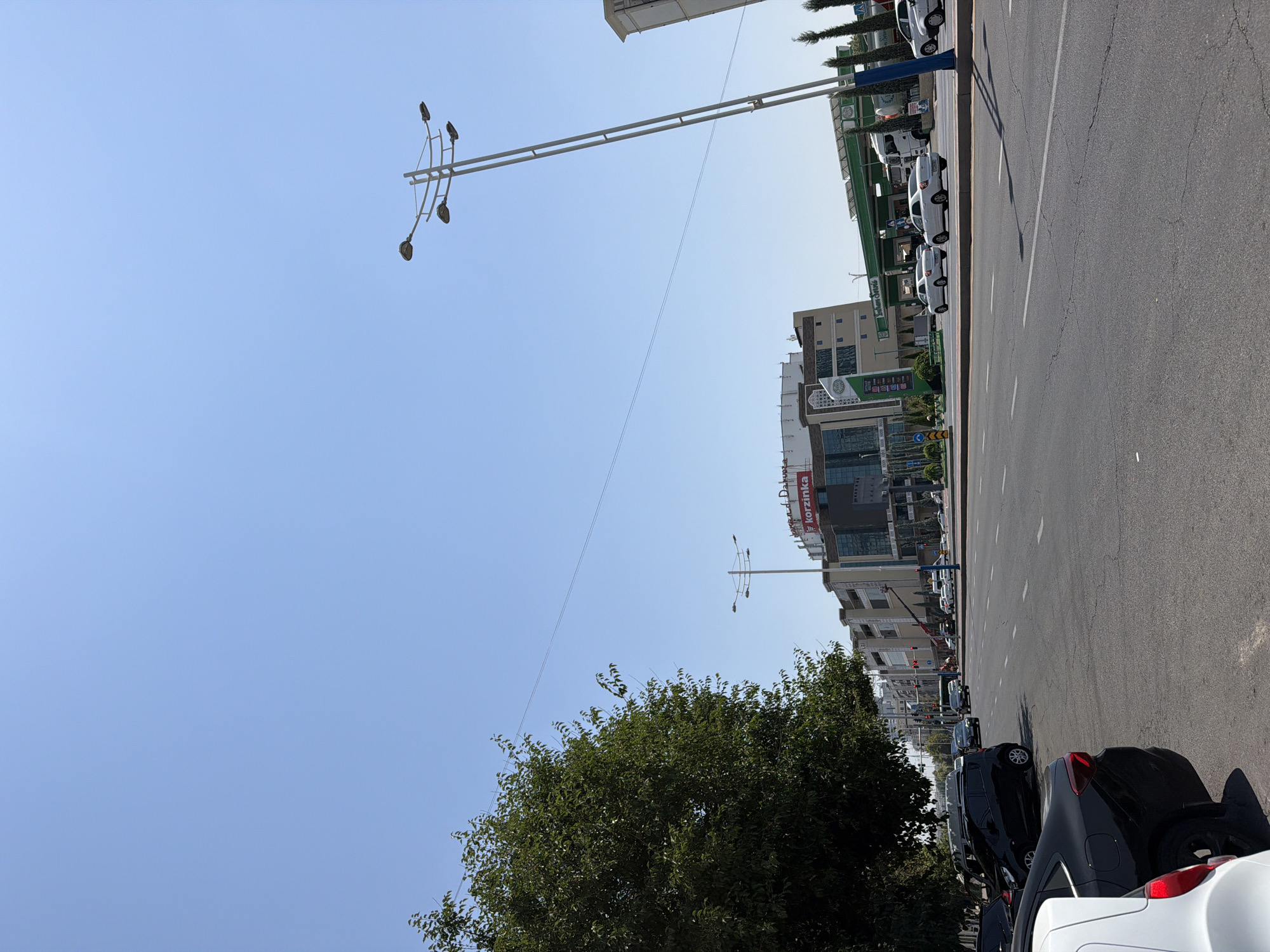 The image captures a street scene in Tashkent, Uzbekistan, on a bright, sunny day with a clear blue sky. In the foreground, the asphalt road is cracked and shows lane markings. Several cars are parked along the left side of the road, including a white car and a black car.
Further down the road, a large tree with dense green foliage stands on the left. Across the street, a modern multi-story building complex is visible. One of the buildings has a large red sign that reads "korzinka". Adjacent to this building is a petrol station with a green canopy and multiple fuel pumps. Several white cars are seen driving on the road. A tall street lamp with multiple lights stands on the right side of the image. The overall impression is of an urban environment with commercial buildings, a main road, and vehicles.
