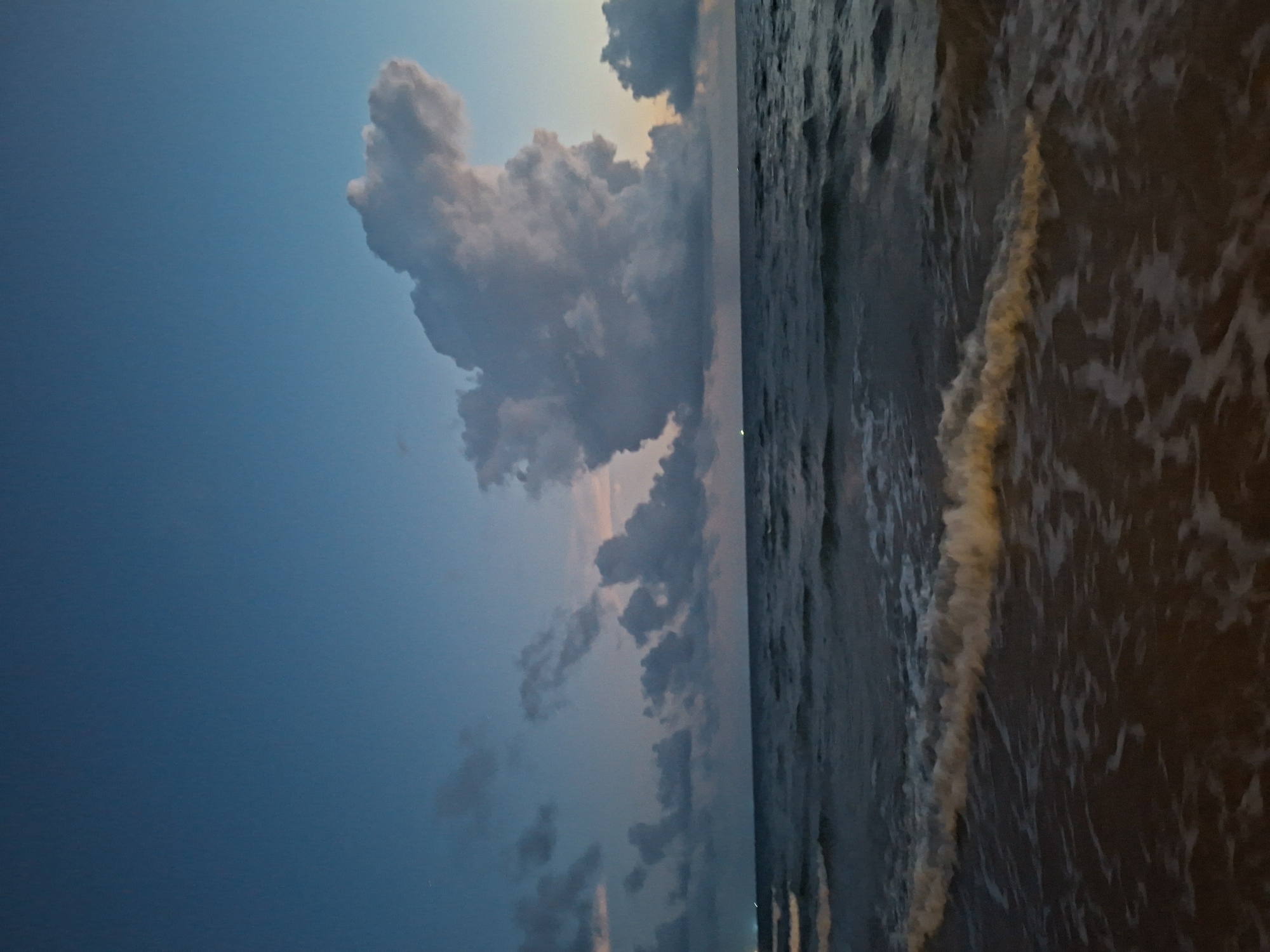 The image depicts a serene seascape at dusk. The sky is a gradient of deep blue at the top, transitioning to lighter shades of blue and hints of pink near the horizon. A large, fluffy cloud formation dominates the upper right quadrant of the sky, catching the last light of the day. Scattered smaller clouds are visible across the horizon. The sea below is calm, with gentle waves rolling towards a sandy shore, which is only partially visible in the foreground. The water is dark, reflecting the twilight sky. A faint glow, possibly from distant lights, can be seen on the horizon line to the left. The overall impression is one of tranquility and the fading light of day. The location is Unknown, Unknown.