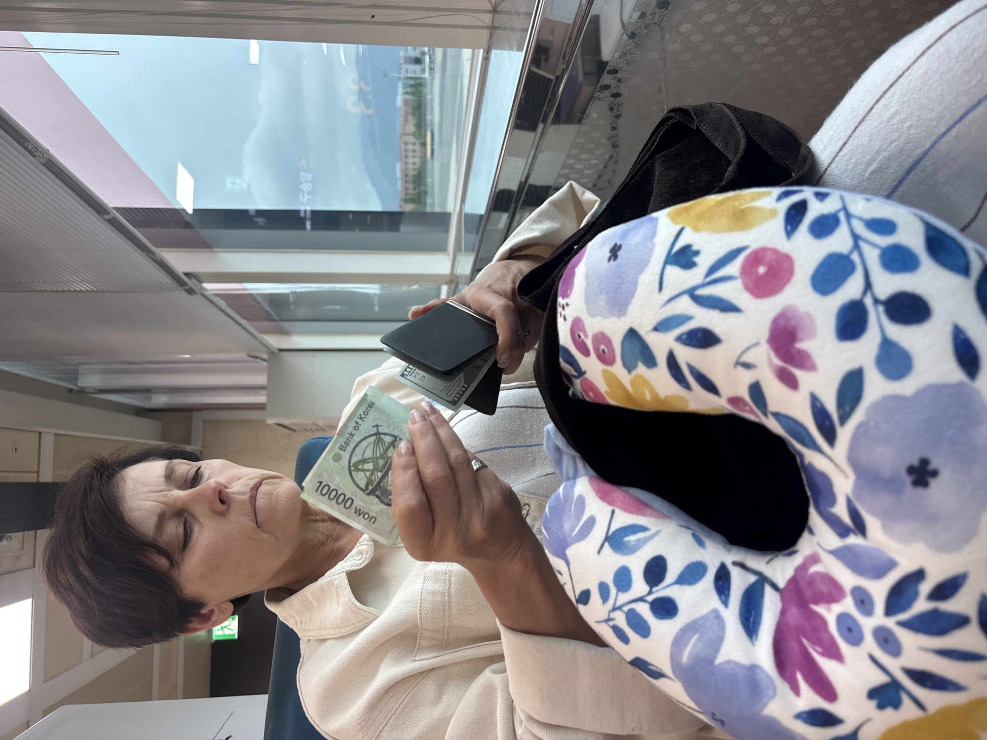 An adult woman with short brown hair is sitting on a train, looking down at a 10,000 Korean Won banknote and a black wallet in her hands. She is wearing a light-colored button-down shirt. In the foreground, a floral-patterned neck pillow is visible. Outside the window, a cityscape with buildings and mountains can be seen under a cloudy sky, suggesting the location is Busan, South Korea.