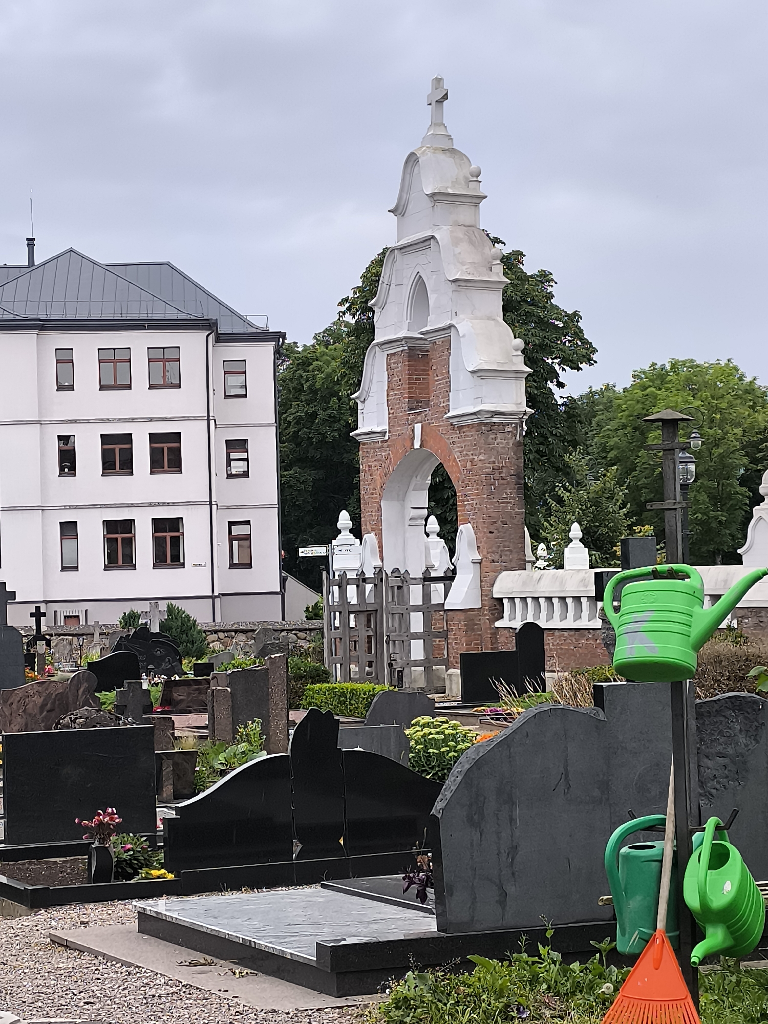 A cemetery scene is depicted in Kretinga, Lithuania under an overcast sky. In the foreground, gravestones of various sizes and materials, predominantly black granite, are visible. Two green watering cans and an orange rake are propped up on a stand to the right of the frame. A white building with a dark gray roof and numerous windows stands behind the cemetery. To the right of the building, a red brick archway topped with white decorative elements and a cross forms part of a structure, possibly a chapel or monument. Lush green trees are present in the background, surrounding the architectural elements and the cemetery. The ground is covered with gravel.