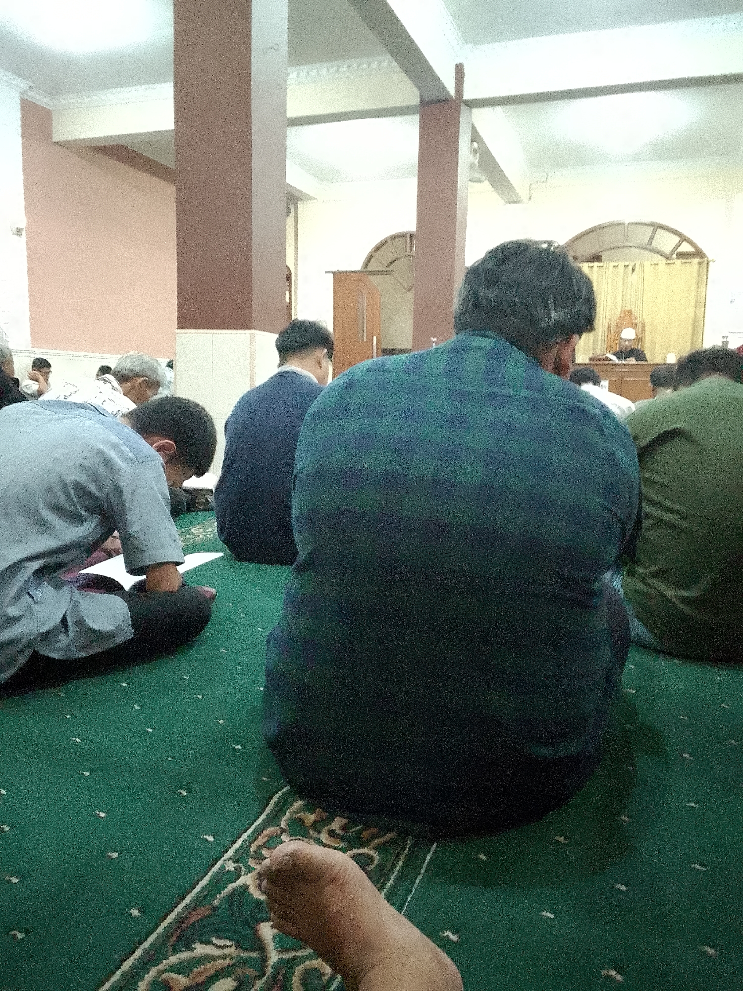 The image depicts an interior scene, likely a place of worship, with several individuals seated on a green carpet. The carpet has a patterned border and small white dots scattered across its surface. In the foreground, the lower leg and foot of a person are visible, with the foot pointing towards the right of the frame.
Several people are seen from behind, with their backs to the camera. One individual in the middle foreground wears a dark blue shirt with a green checkered pattern. To their left, a person in a light blue garment is seated and appears to be looking down at something in front of them, possibly a book. To the right of the person in the checkered shirt, another individual wears a dark blue shirt. Further to the right, a person in a green shirt is visible. In the background, more individuals are seated, and some appear to be looking towards a raised platform or lectern where a person in a white hat and dark clothing is seated. The architecture includes large, square columns with reddish-brown exteriors and light-colored ceilings. Arch-shaped window frames or decorative elements are visible in the background, covered by light-colored curtains.
The lighting suggests it is daytime, with the light coming from above and through the background elements. The scene is static and appears to be a moment of quiet activity, possibly prayer or a religious lecture. Without further context, the specific city of Kertajaya, Indonesia, cannot be definitively identified from the visual cues alone.