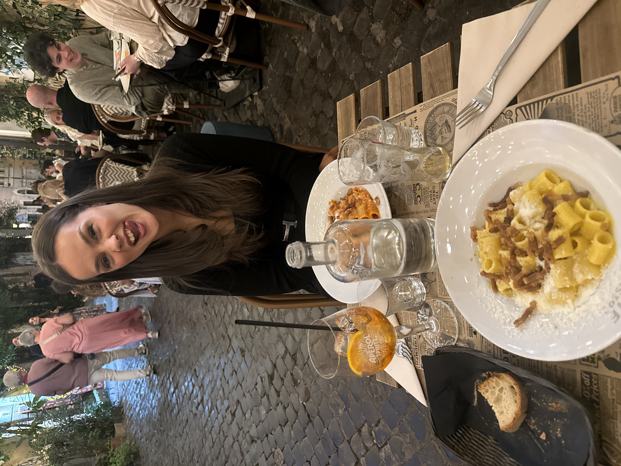 The scene captures an outdoor dining setting in Rome, Italy, characterized by a cobblestone street and tables arranged on the sidewalk. A young woman with long brown hair, wearing a black top, is the primary subject, smiling at the camera. On the table in front of her, there is a plate of pasta with sauce, a plate of carbonara pasta, a water bottle, several glasses, an Aperol Spritz, and a piece of bread in a black bowl. The table is covered with what seems to be a newspaper-themed tablecloth. In the background, other people are walking on the sidewalk and dining at other tables. The setting indicates a typical Roman street with trees and buildings blurred in the background.