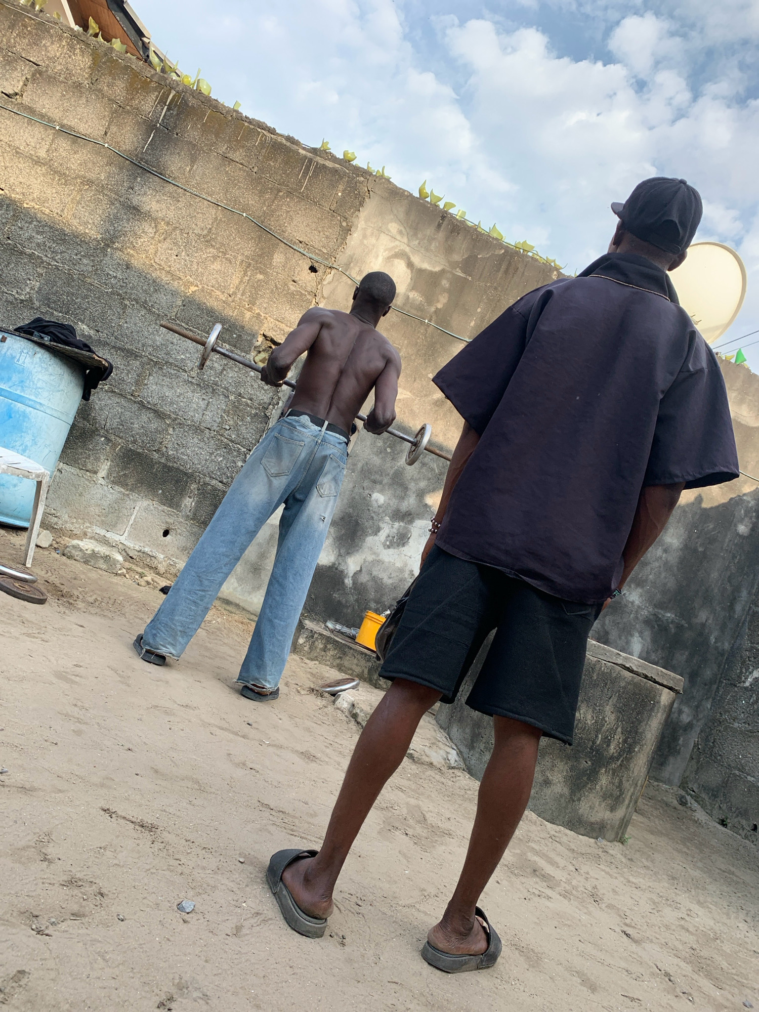 The image depicts two individuals in an outdoor urban setting, likely in Ado, Nigeria, characterized by a rough concrete wall forming the backdrop, partially obscured by vegetation at the top. The ground is composed of sandy soil.
The individual in the foreground is facing away from the camera, wearing a dark t-shirt and black shorts. They are also wearing dark sandals on their feet.
The individual in the mid-ground, also facing away from the camera, is shirtless and appears to be engaged in weightlifting with a barbell. They are wearing light-wash denim jeans and dark sandals. Their physique suggests they are muscular.
Several objects are visible in the scene: a blue cylindrical container is positioned to the left of the frame, a white plastic chair is situated near the container, and weight plates are scattered on the ground near the chair and to the right of the image. A large, rough concrete structure is visible to the right of the frame. The sky above is partly cloudy.