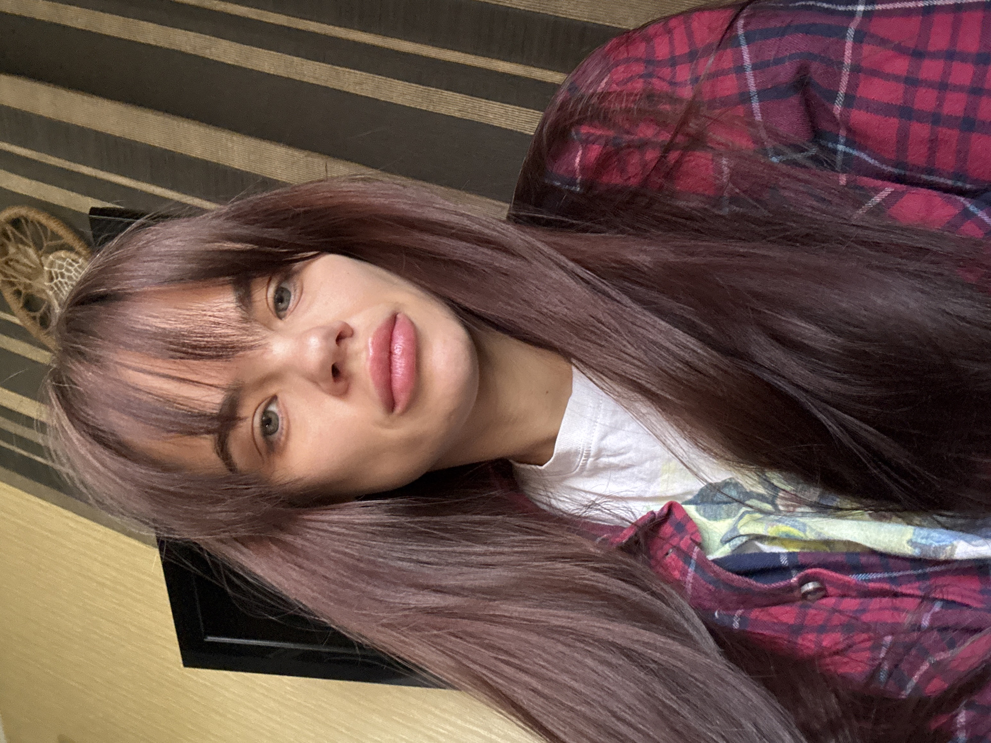 A woman with long, straight brown hair and bangs is posing for a selfie. She is wearing a red and black plaid shirt over a white t-shirt. The background includes a wall with brown and beige vertical stripes, a dreamcatcher hanging on the wall, and a black, flat-screen television. There is nothing in the image to indicate a location in the city of Rostov-on-Don, Russia.