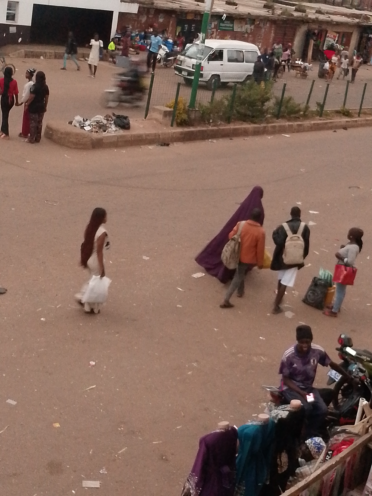 The image shows a street scene in Bukuru, Nigeria. In the foreground, a person wearing a purple t-shirt and a dark beanie sits on a motorcycle. The motorcycle is dark in color and has a visible exhaust pipe and rear wheel. To the left of the motorcycle, several mannequins display clothing, including a purple garment and a teal garment. The mannequins are arranged on a wooden structure, which appears to be a stall or display. The ground is covered in dirt and debris, with some scattered papers and plastic.
In the midground, several people are walking on the street. A person in a light-colored garment with dark hair walks away from the camera, carrying a white plastic bag. Further back, two individuals are walking towards the camera. One is wearing an orange jacket and carrying a bag over their shoulder. The other person is wearing a dark jacket and a backpack, with white shorts. Behind them, a figure in a dark purple, hooded garment walks.
In the background, several buildings and a white van are visible. The buildings appear to be shops or residences. Some people are seen near the buildings, and a white van is parked on the street. The overall environment suggests a busy urban setting, likely a market or commercial area. The lighting indicates it is daytime.