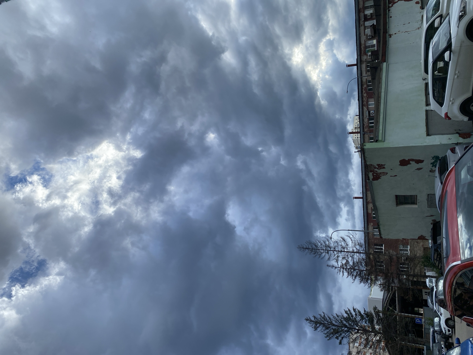 A wide-angle, outdoor, eye-level shot depicts a cloudy, possibly overcast sky above an urban scene in Novokuybyshevsk, Russia. The sky is filled with dramatic, dark grey clouds with patches of lighter, possibly sunlit clouds breaking through in the upper central portion. Below the sky, a row of buildings is visible. On the left, a tall residential building is partially obscured by two tall, bare evergreen trees. Adjacent to these trees is a longer, lower industrial-style building with brickwork and visible windows. In front of this building, a pale green, dilapidated structure with peeling paint is prominent. The foreground is occupied by a parking lot filled with various cars, including a red car in the lower left corner, a white SUV on the right, and other vehicles partially visible. The scene appears to be set during daytime, but the heavy cloud cover suggests a subdued lighting condition, potentially indicating an approaching storm or a change in weather. No people are visible in the frame.