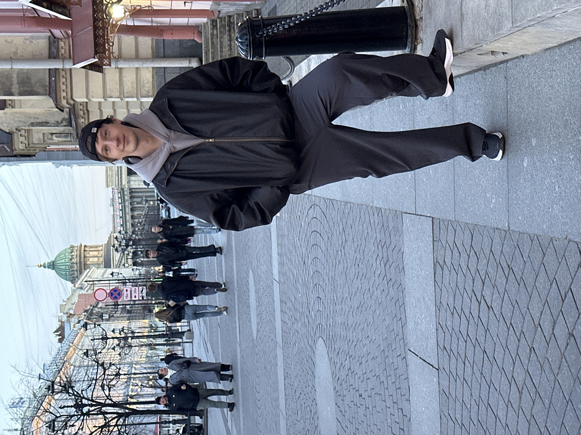 The image depicts a man standing on a paved area in what appears to be a city street. He's wearing a black jacket, a gray hoodie, dark pants, and a cap. He is looking directly at the camera.
Behind him, a line of pedestrians walks along the same street. In the background, the dome of Kazan Cathedral is visible. Streetlights line the street. The location appears to be in Saint Petersburg, Russia. The pavement is made of small square stones arranged in a circular pattern.