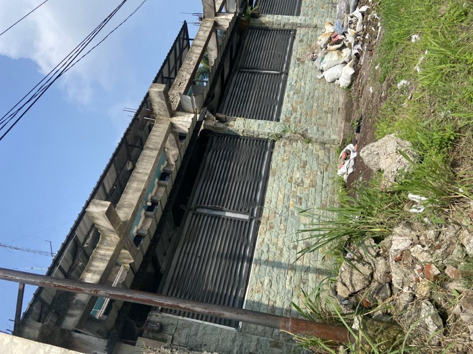 The image shows a concrete wall with a black metal fence and a stone facade. The wall is located on a sloping lot, and the ground is covered in grass and rubble. There are several white bags and other debris lying on the ground. The scene is taken from a low angle, looking up at the wall. The coordinates provided indicate a location in the city of Margaasih, Indonesia, which is about 10 kilometers south of Bandung, the capital of West Java.