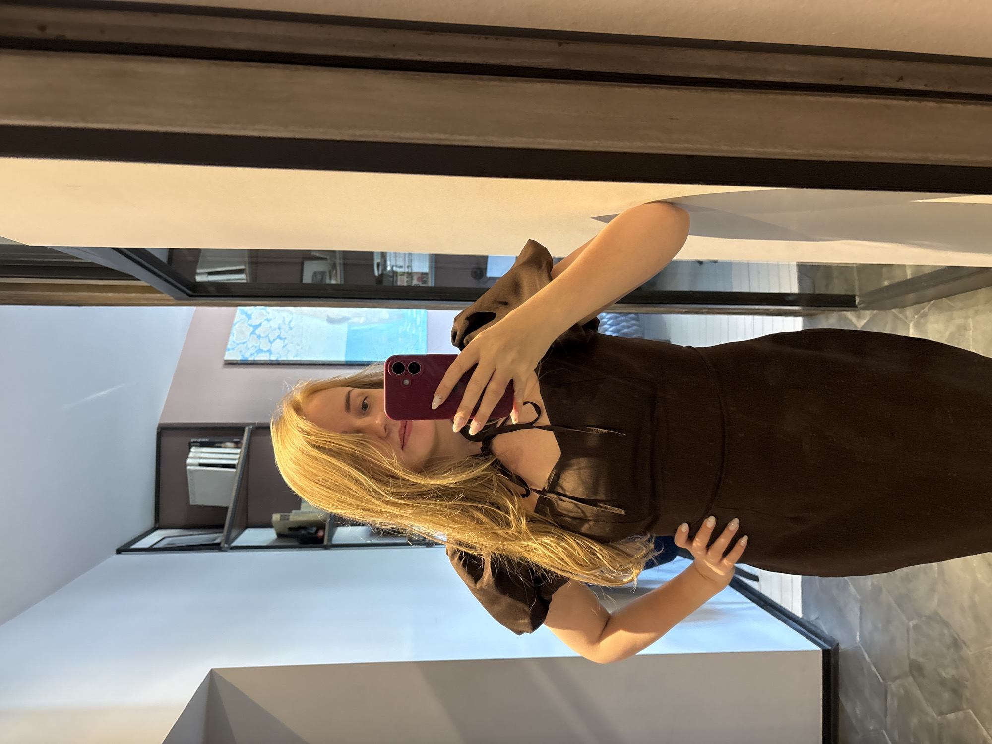 A woman with long blonde hair is taking a selfie in what appears to be an indoor setting. She is holding a dark-colored smartphone and wearing a dark brown dress with puff sleeves and a tie detail at the chest. She is standing in front of a mirror. The background includes elements such as shelves with objects on them and a glimpse of what might be an outdoor scene with blue hues, possibly water and sky. The scene lacks distinctive landmarks and precise details, making it impossible to confirm if it is in Saint Petersburg, Russia, or to pinpoint a specific location.