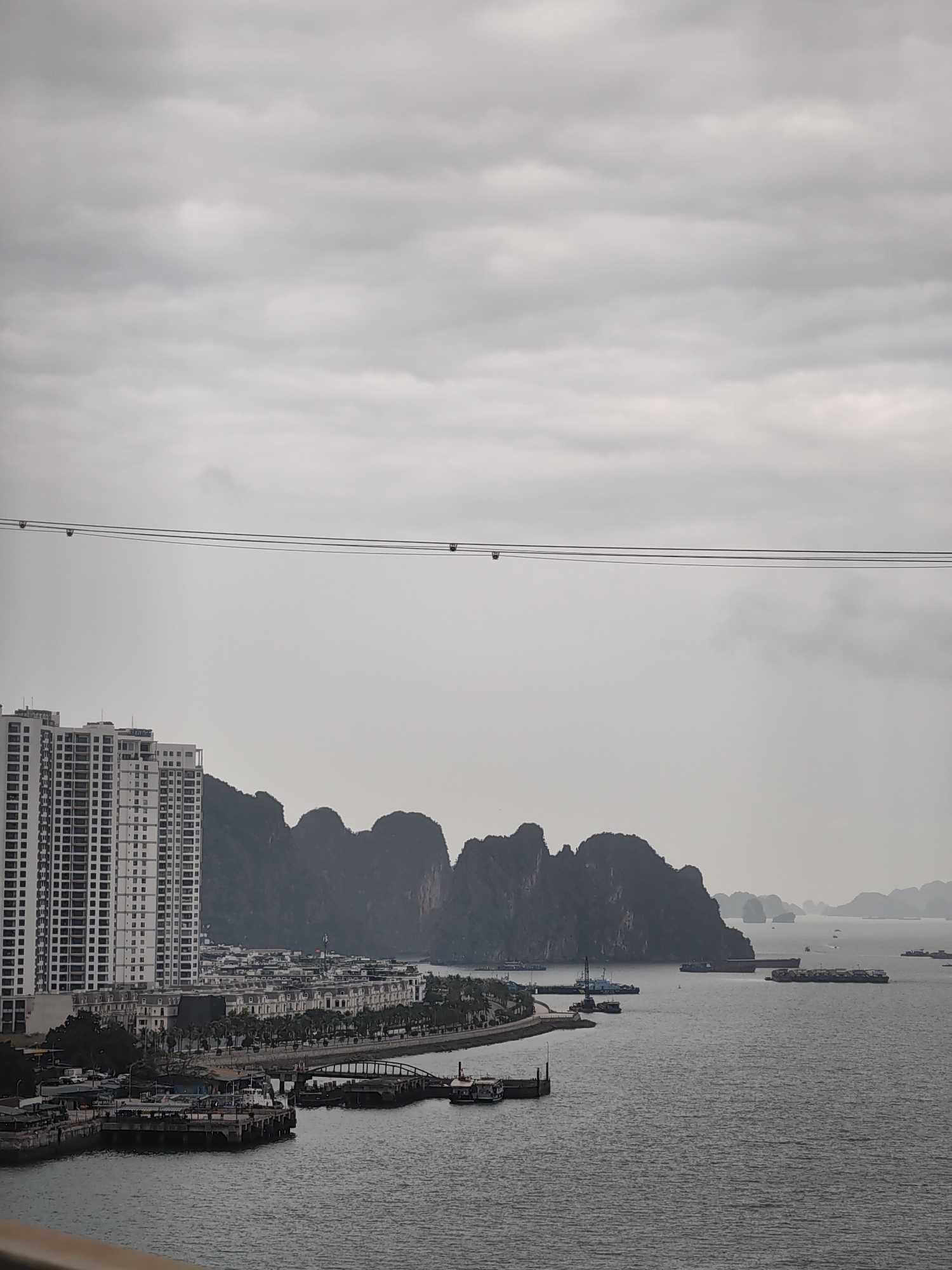 A high-angle view shows Ha Long Bay in Bai Chay Ward, Vietnam, on an overcast day. Tall, modern apartment buildings line the coast, with a row of townhouses behind a line of palm trees. There are piers and docks, with various boats at anchor. The background features karst formations rising from the water. A cable-car line is in the sky.
