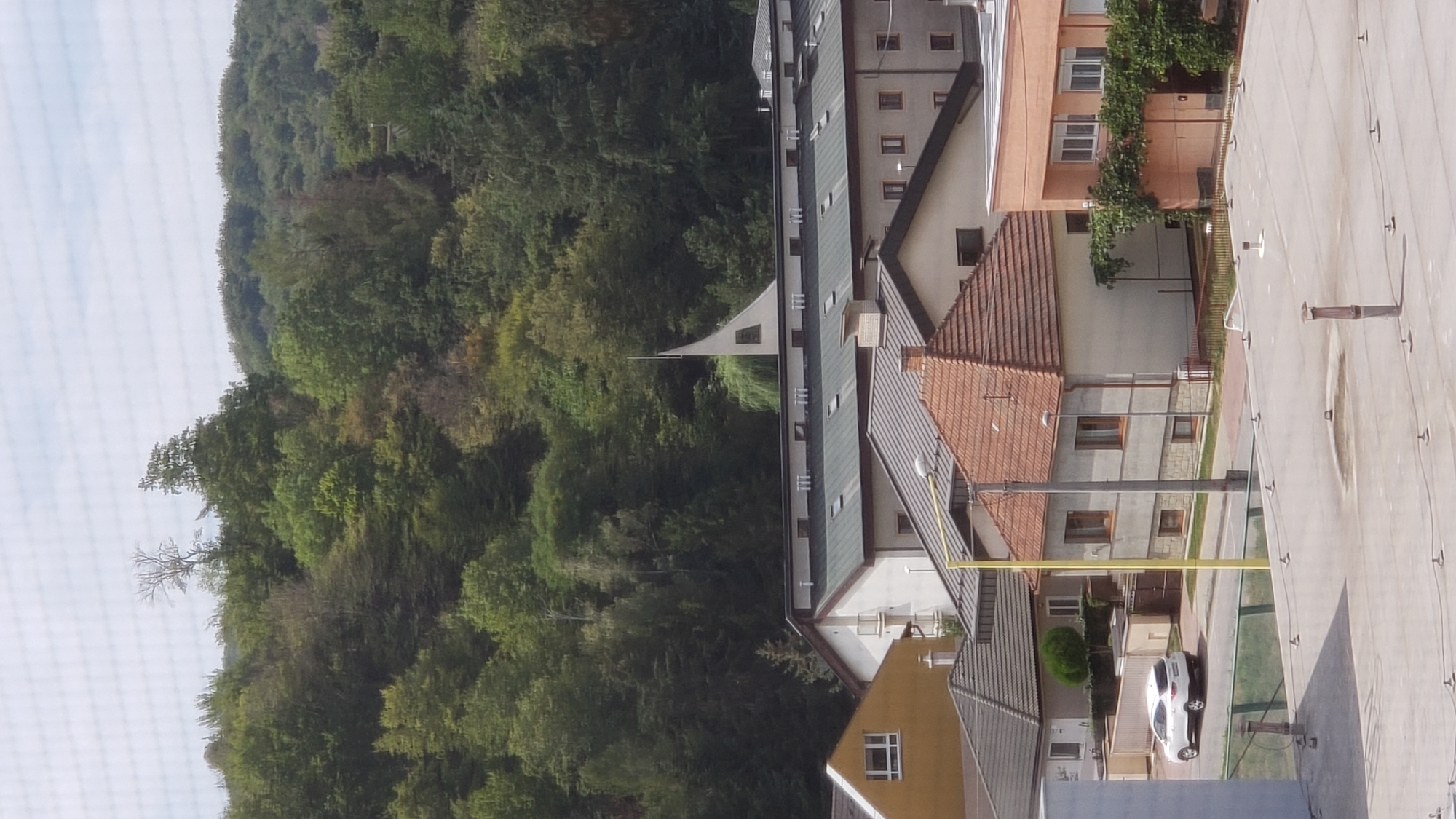 The image depicts an aerial view of a residential area with a wooded hillside in the background, likely located in Prešov, Slovakia. In the foreground, several rooftops are visible, some with peaked tile roofs and others flat. A white SUV is parked on a street in front of a low-rise building with a yellow facade. Further back, a large building with a dark, sloped roof features numerous skylights. A distinctive white structure with a pointed, sail-like top rises above this building. The wooded hillside is densely covered with green trees, with a few bare branches visible towards the top. The sky is partly cloudy.
