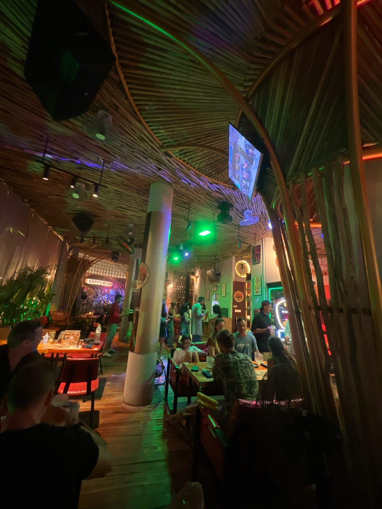 The image depicts the interior of a restaurant or bar, possibly "Happy Bear" as indicated by a sign. The architecture features a distinctive ceiling made of natural materials, resembling bamboo or wood, with exposed beams and framing. The space is lit with a combination of ambient and colored lighting, including green and purple hues, creating a vibrant atmosphere. Several patrons are seated at tables and standing, engaged in conversations and enjoying their time. The overall scene suggests a casual and lively environment, possibly a tourist hotspot in the city of Phú Quốc, Vietnam, known for its tropical ambiance and entertainment venues.