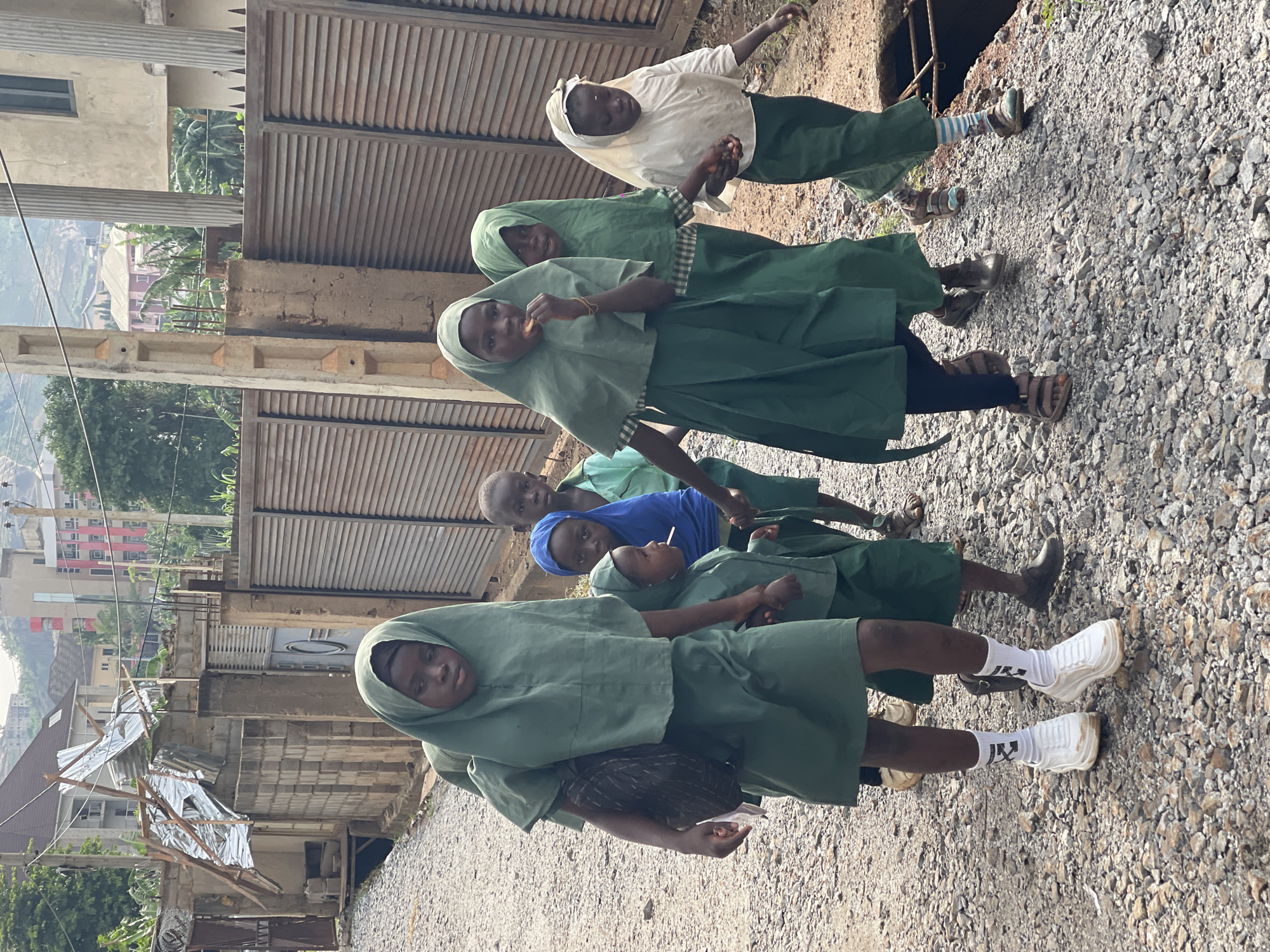 In Dutsen Alhaji, Nigeria, a group of six children stand on a rocky, gravelly road near a concrete structure and a corrugated metal gate. Five of the children wear green school uniforms with headscarves, and one child wears a blue shirt. One child near the front carries a dark-colored bag. The background includes buildings, a partially constructed or damaged structure, and a hillside.
