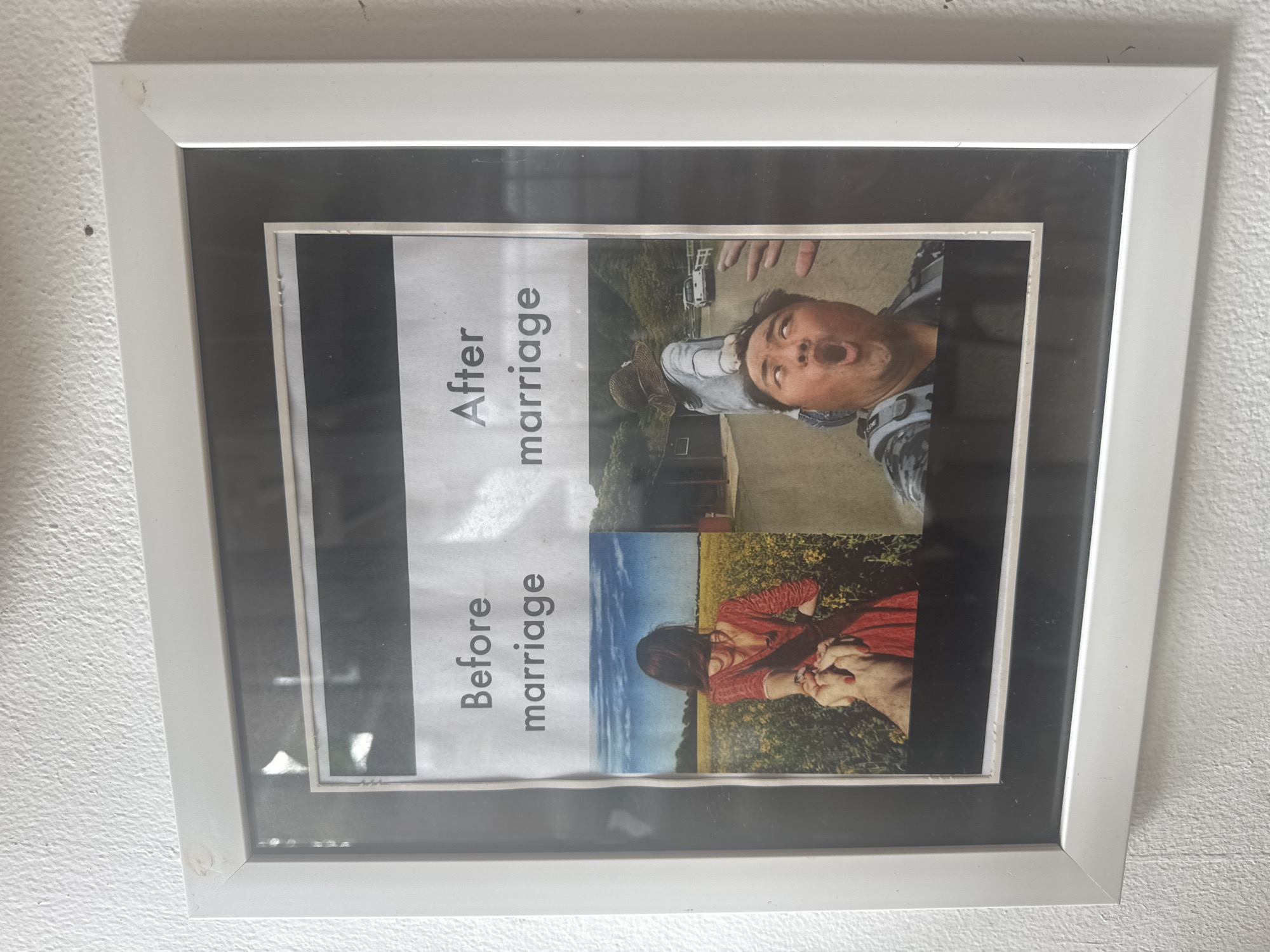 A framed image hangs on a white wall, depicting a before-and-after marriage meme. The frame is white with a black inner border. "Before marriage" is printed in bold text above an image of a couple holding hands, appearing to be in a field. "After marriage" is printed above an image of a man with his mouth open and eyes wide in a comical expression, appearing to be outdoors. The image is not taken in Nusa Penida, Indonesia.