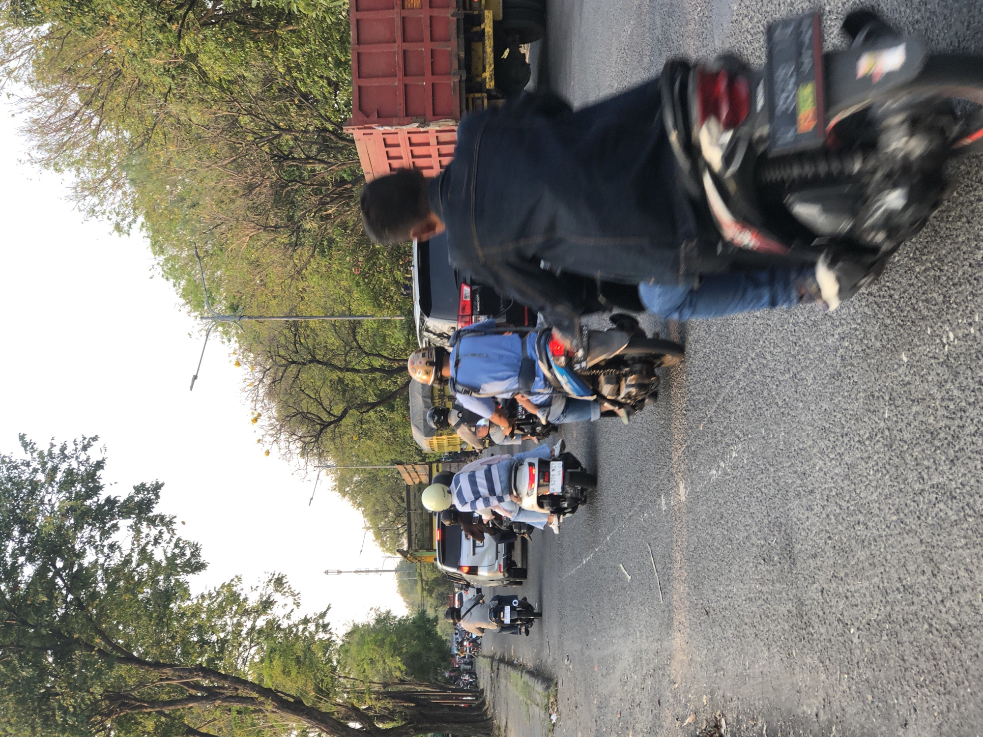 In Gelam, Indonesia, a street scene unfolds with multiple motorcycles navigating a road alongside cars and trucks. A man in a denim jacket rides a motorcycle in the foreground, his back to the camera. Behind him, another rider in a blue shirt and jeans also rides a motorcycle. Further ahead, a motorcycle carries two people, one wearing a striped shirt and a light-colored helmet, the other a darker helmet. A white SUV is visible in the lane to the left, followed by a black SUV. A large red truck with a yellow chassis is to the right. Trees line the road on both sides, with power lines and streetlights interspersed. The road surface is asphalt. The sky is overcast.