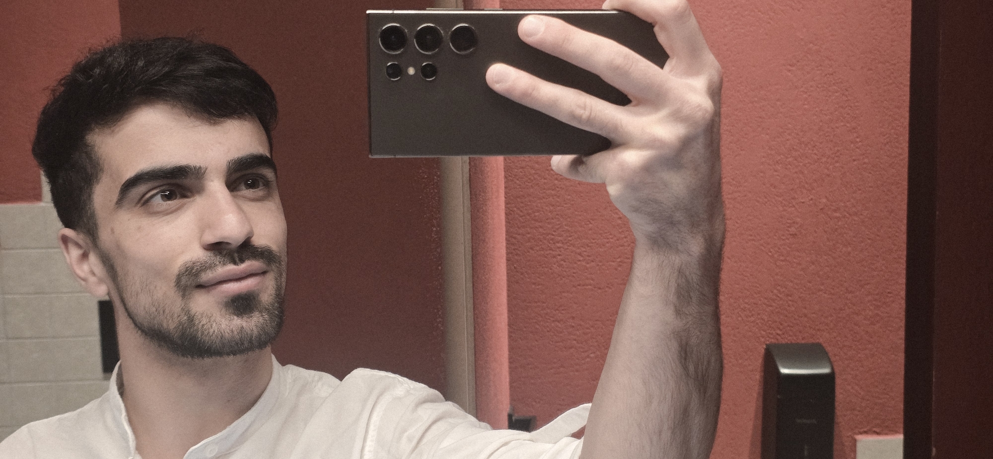 A man with dark hair and a beard is taking a selfie with a black smartphone. He is wearing a white shirt. The background consists of a red wall and a tiled wall in the lower left corner. A black object is visible on the right side of the image. The location in Yerevan, Armenia, cannot be determined from the image.