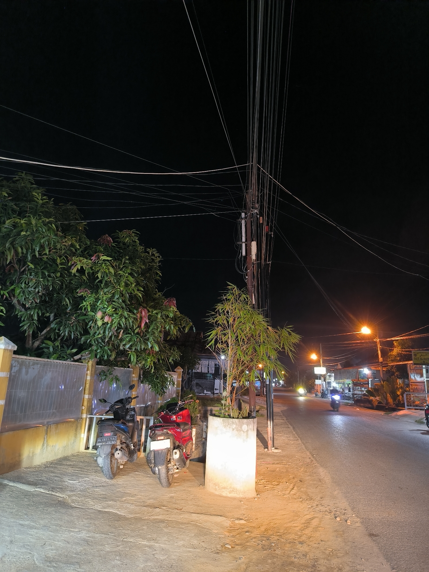 The image depicts a street scene at night in Lhokseumawe, Indonesia. A road curves to the right, illuminated by streetlights and the headlights of passing vehicles, including a motorbike in motion on the right side of the road. On the left side of the road, two parked motorcycles are visible, one red and one blue, in front of a yellow-painted wall and a translucent fence. Tall trees with abundant foliage, some bearing fruit, are situated behind the fence. A concrete planter containing bamboo plants stands on the sidewalk. Overhead, a dense network of electrical wires crisscrosses the dark sky, supported by a utility pole. Several buildings with illuminated signs and open structures line the right side of the street, suggesting commercial establishments. The overall ambiance is that of a well-lit urban street at night.