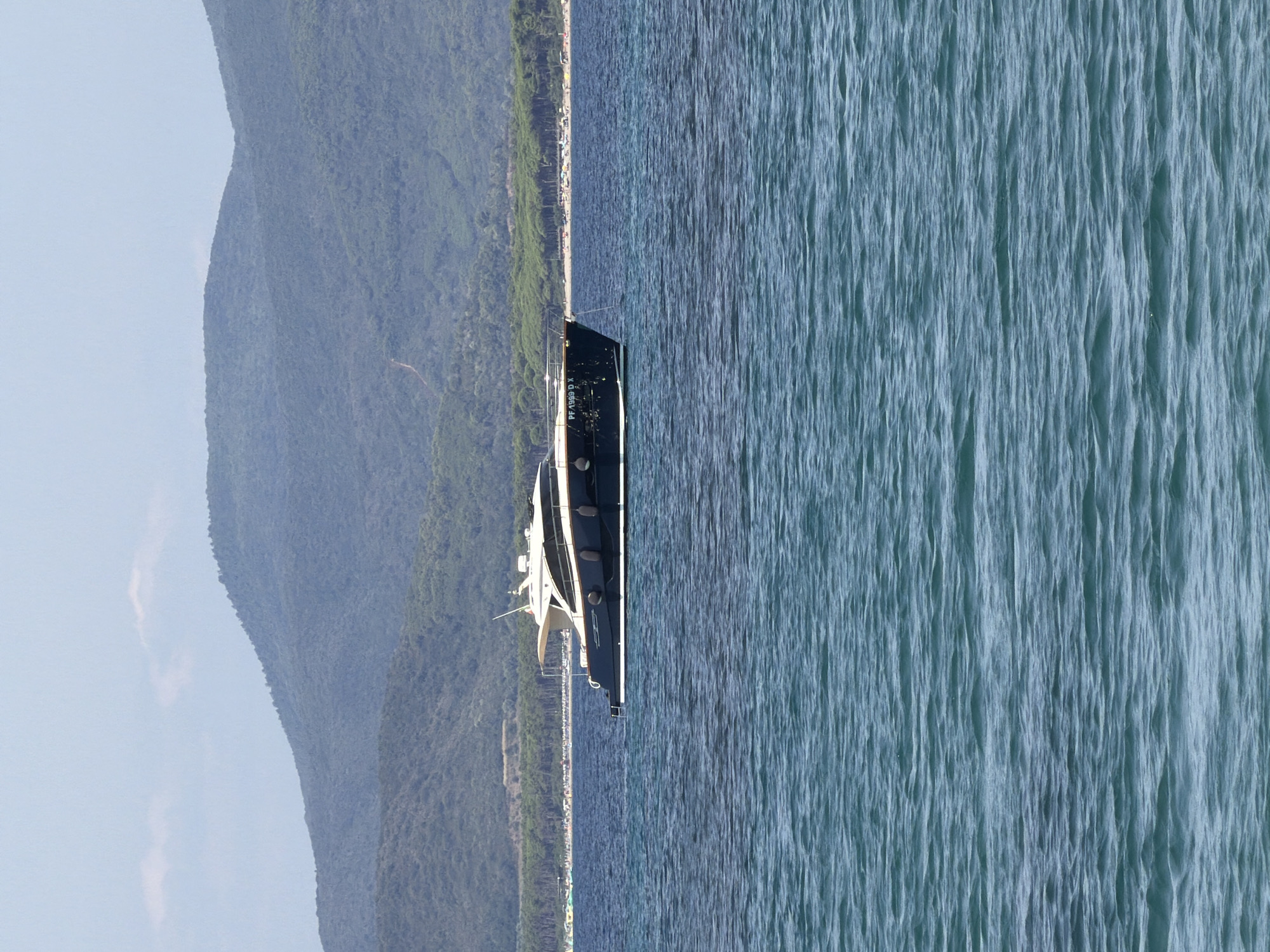 A dark blue yacht with a white top is anchored in the choppy, turquoise waters of the Mediterranean Sea. The yacht has a registration number "PF 1909 D X" visible on its bow. In the background, a tree-lined beach with numerous sunbathers and umbrellas stretches along the shore. Behind the beach, rolling, green hills covered in dense foliage rise under a hazy, light blue sky with a few wispy clouds. The scene suggests a serene summer day in Punta Ala, Italy.