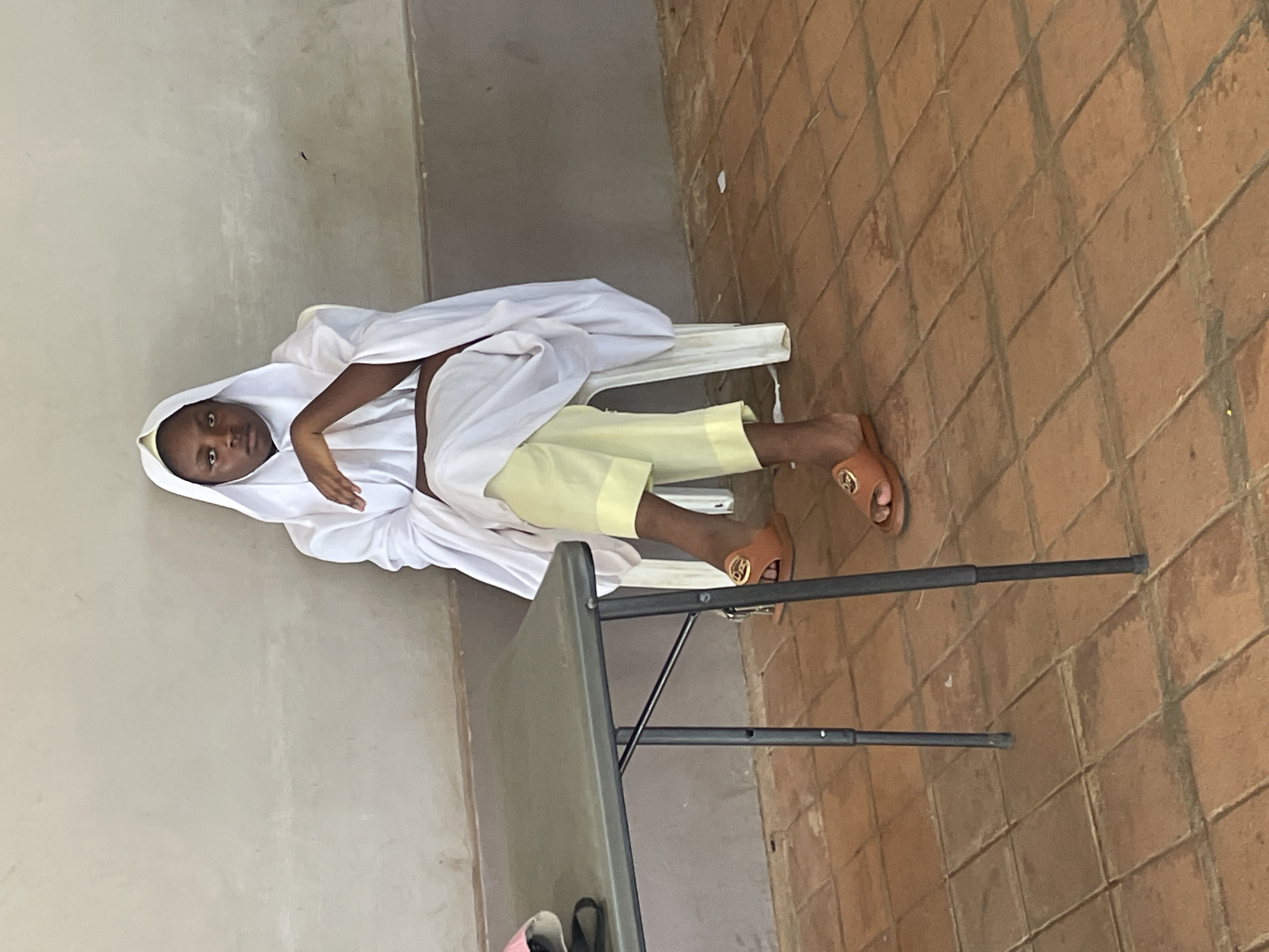In Barakin Lerawi, Nigeria, a young Black female sits on a white plastic chair, facing forward, in front of a light grey concrete wall. She wears a white headscarf and a long, flowing white garment over pale yellow trousers. Her right arm rests on her lap, while her left arm is bent and her hand is positioned on her chest. She wears brown slide sandals with gold buckles. To her left, a dark grey metal folding table with a textured surface is positioned. The floor is tiled with reddish-brown terracotta squares. The overall environment appears to be outdoors or in a covered outdoor space.