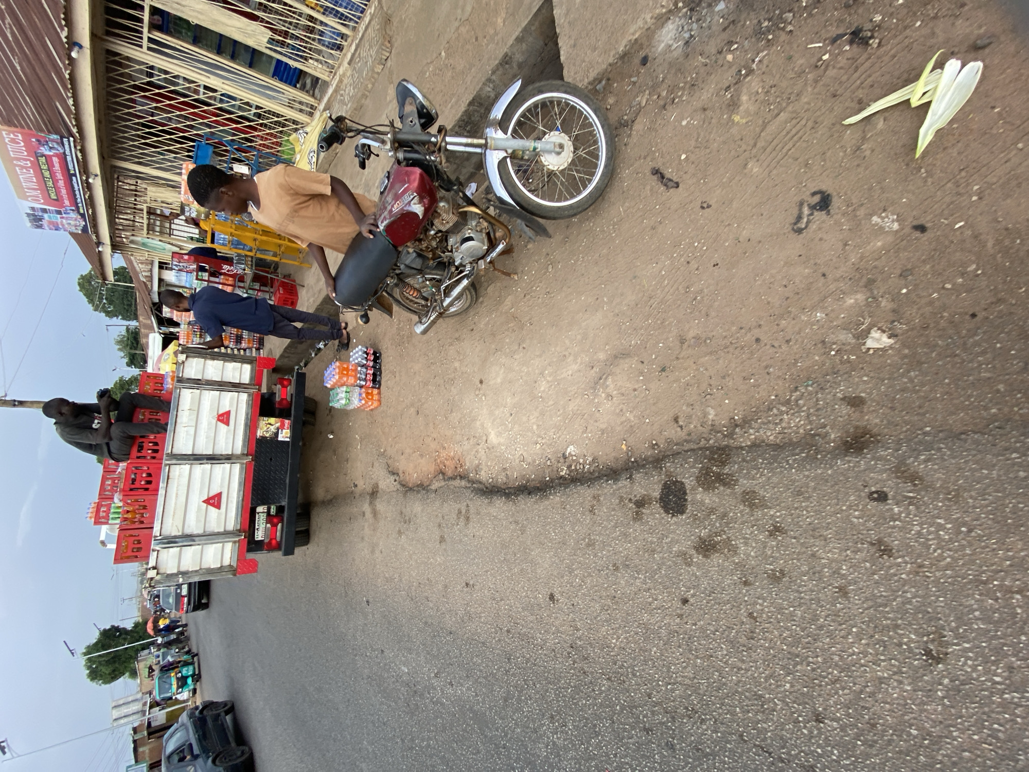 This image captures a vibrant street scene in what appears to be a commercial area in Jos, Nigeria. The perspective is slightly overhead, looking down at a street with a dirt and concrete shoulder.
In the foreground, a man in a tan shirt and dark pants stands beside a red and silver Bajaj motorcycle, which is parked on the dirt section. His posture suggests he might be adjusting something or preparing for departure. Further down, another man in a dark blue shirt is seen near a white and red truck. The truck is heavily laden with stacks of red and green plastic crates, predominantly filled with bottled soft drinks like Coca-Cola, Fanta, and Sprite. More stacks of these bottled beverages are also arranged on the ground beside the truck. A third man, dressed in dark attire, is seated on top of some red crates on the rear of the truck, facing away from the camera.
Adjacent to the sidewalk is a building with a corrugated metal roof and white security grilles, indicating a shop. A prominent sign above this shop reads "O.M. WINE & JUICE WHOLE SALE AND RETAIL," with smaller text beneath listing "DRINKS, BEVERAGES, PROVISIONS, COSMETICS, ETC."
The street itself shows signs of activity, with other vehicles like cars and possibly tricycles visible further down the road, suggesting a bustling environment. The ground on the shoulder is a mix of dirt and concrete, with some scattered debris and leaves. The truck's license plate, partially visible, reads "JJT 516 X ABUJA," confirming a Nigerian setting. The daylight suggests it is either morning or afternoon, with no harsh shadows indicating midday sun. The scene depicts a typical commercial exchange or delivery operation in a busy urban setting.