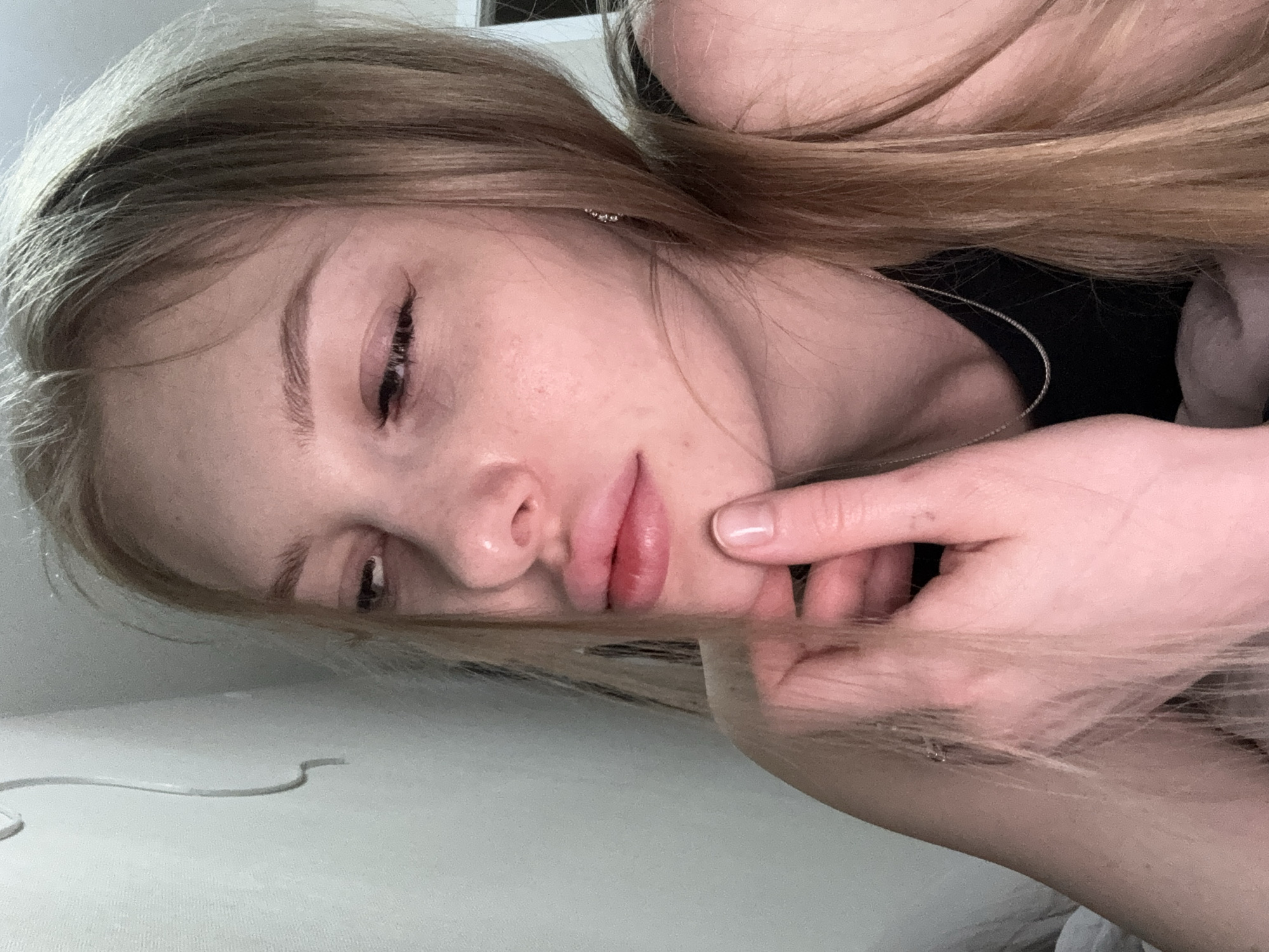 A fair-skinned individual with blonde hair is lying down. Their left hand is touching their face. The background is a light color. A cord is visible on the left side. A necklace is around the person's neck. Location unknown, but given the prompt, possibly Perm, Russia.